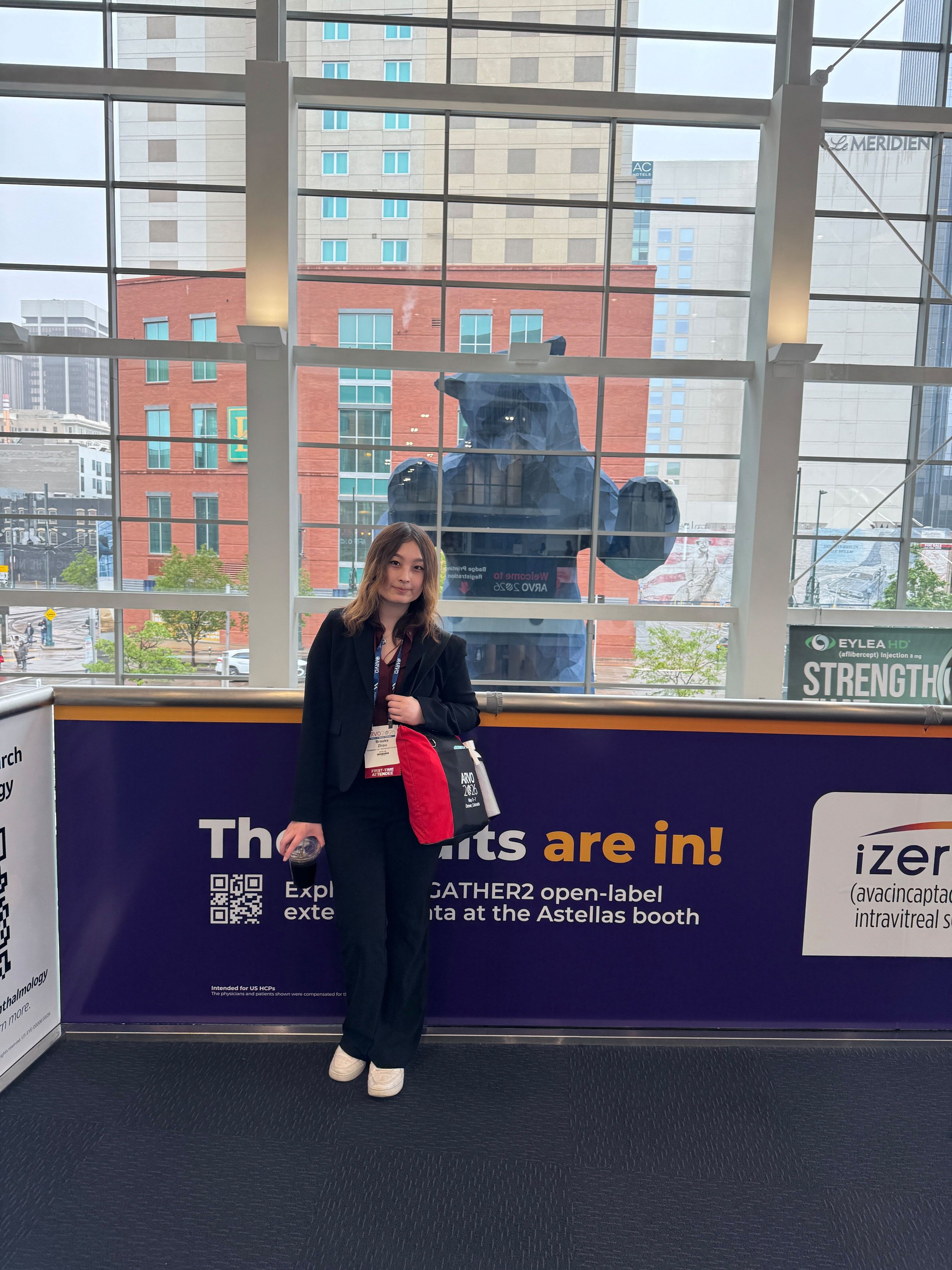 A woman standing in front of a conference poster at an indoor event. Behind her is a large window with a cityscape view, including buildings and a bear mascot outside the window.