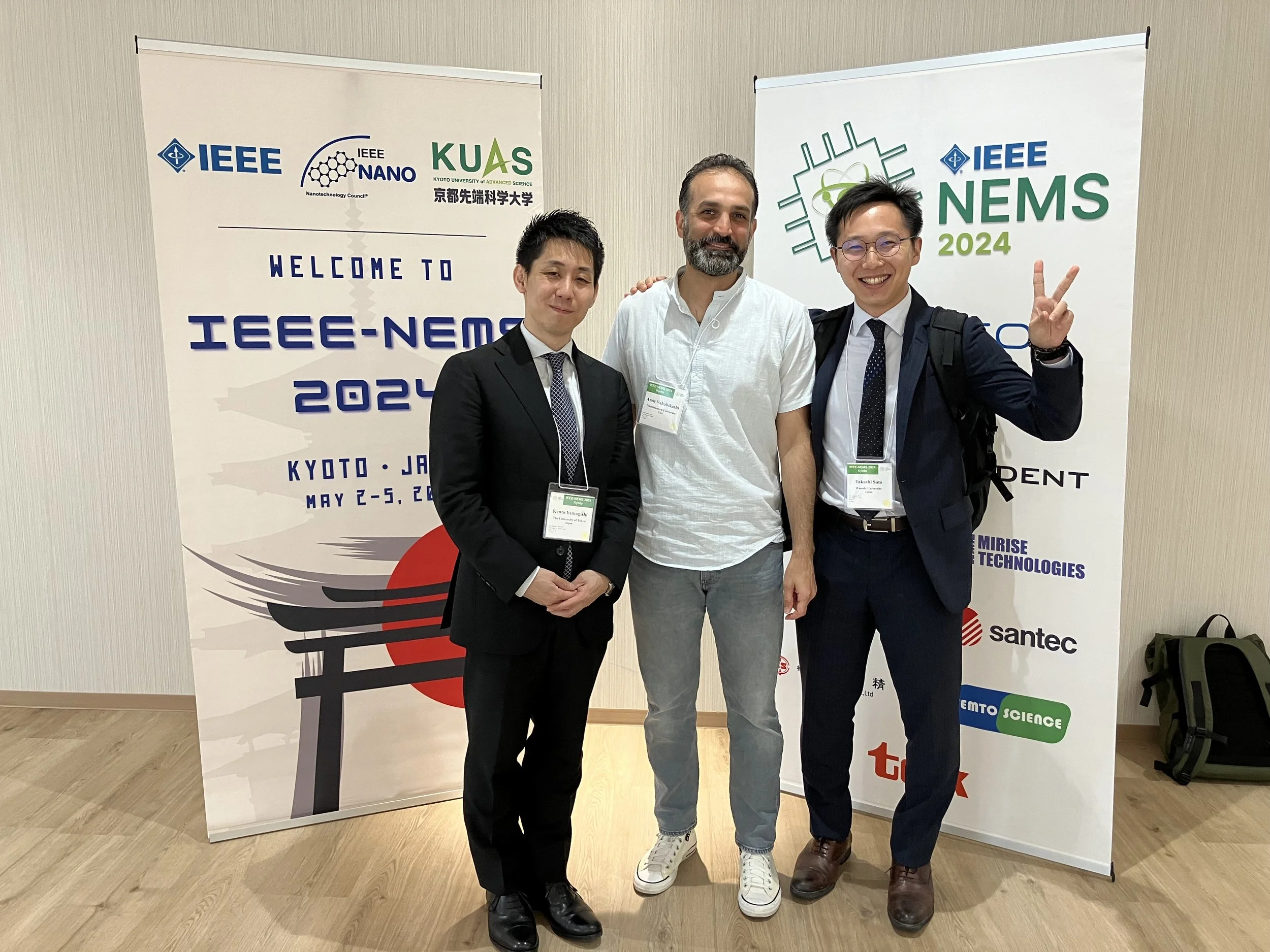 Three men standing together in front of two banners at an academic conference. The man on the left is wearing a black suit and tie, the man in the middle is wearing a white shirt, and the man on the right is wearing glasses, a dark suit, and a tie, making a peace sign with his hand.