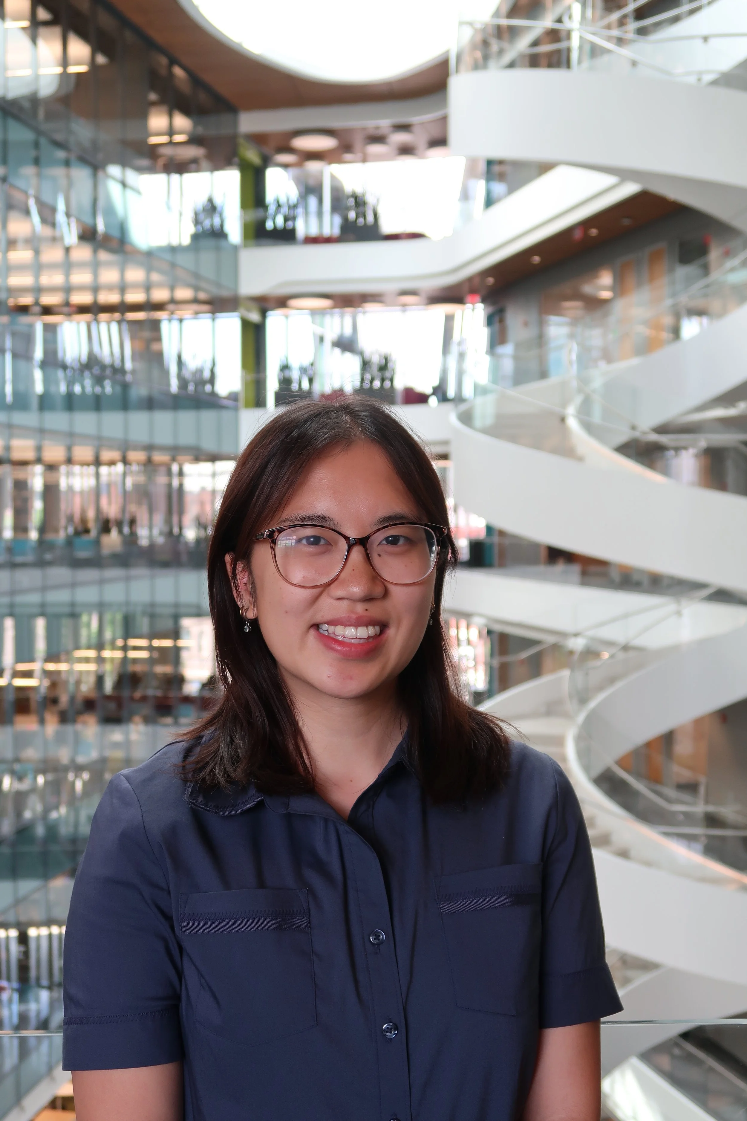A woman with glasses and long dark hair smiling at the camera inside a modern building with a spiral staircase and glass walls.