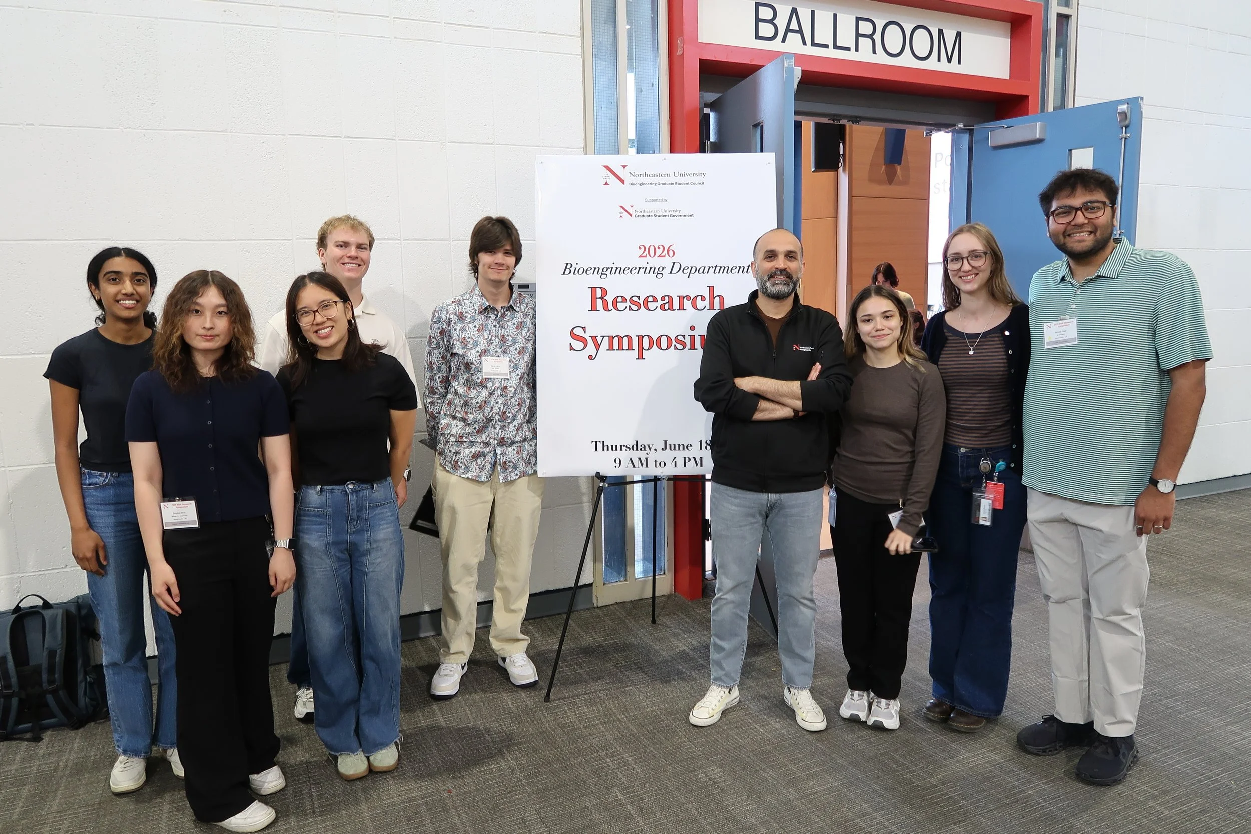 Group of nine diverse young adults standing in a line at a research symposium, smiling, outside a ballroom entrance, with a white sign displaying event details.