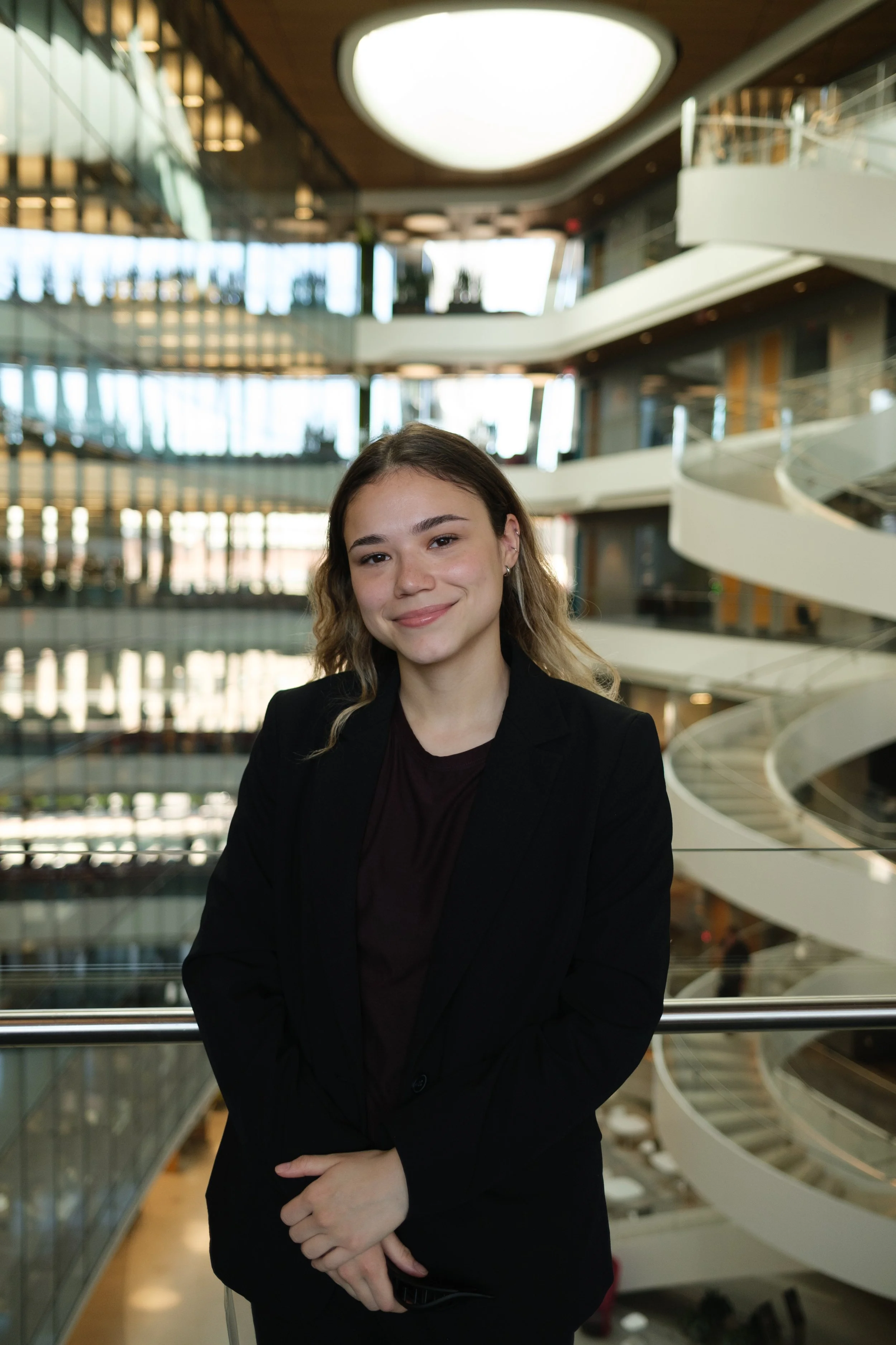 A young woman with shoulder-length wavy hair, wearing a black blazer and dark top, standing in a modern building with multiple floors, glass railings, and an architectural spiral staircase in the background.