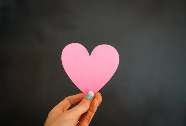 A hand holding out a pink heart paper cut out.