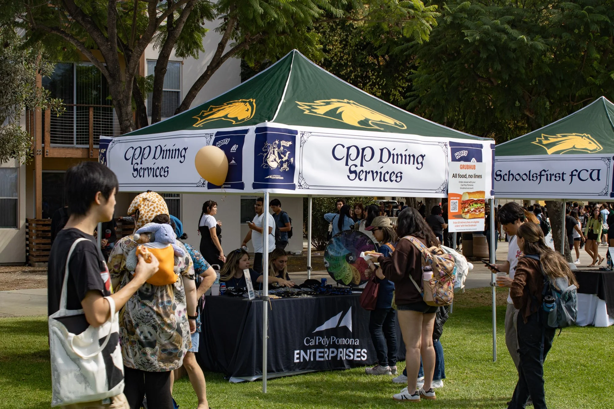 A outdoor event with multiple tents and gathering of people, some of whom are standing and sitting near tables. One prominent tent displays the logo and name of 'Cal Poly Pomona Enterprises' and 'CPP Dining Services'. There are trees and a building in the background, and a person is holding a taco in the foreground.