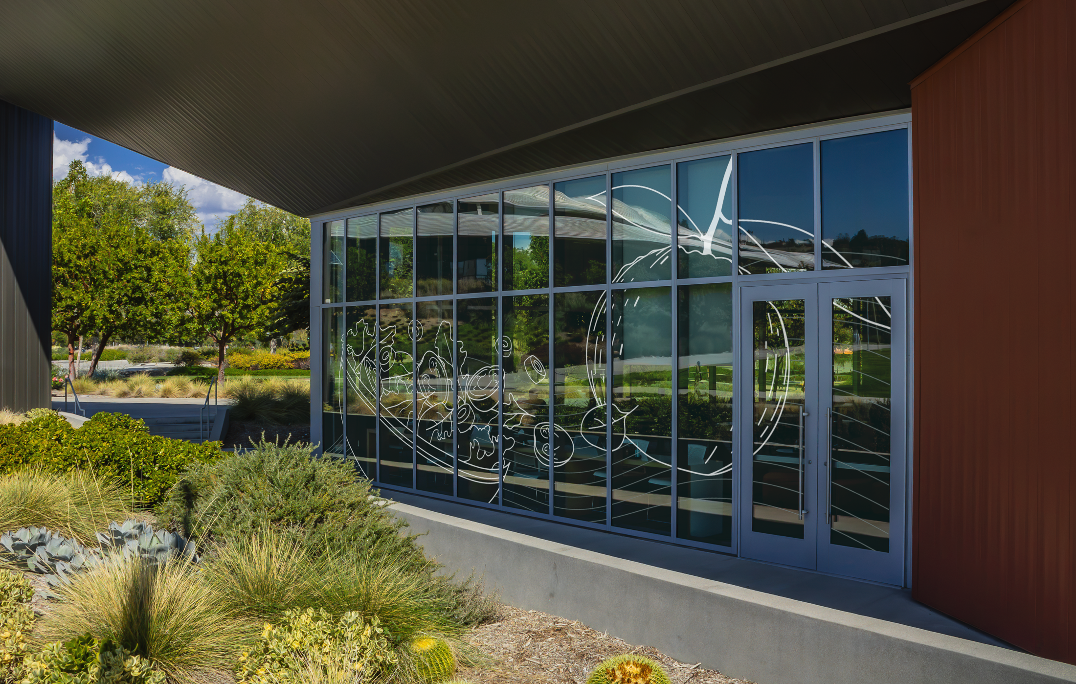 Modern building with large glass windows reflecting trees and a blue sky, with desert plants in the foreground.