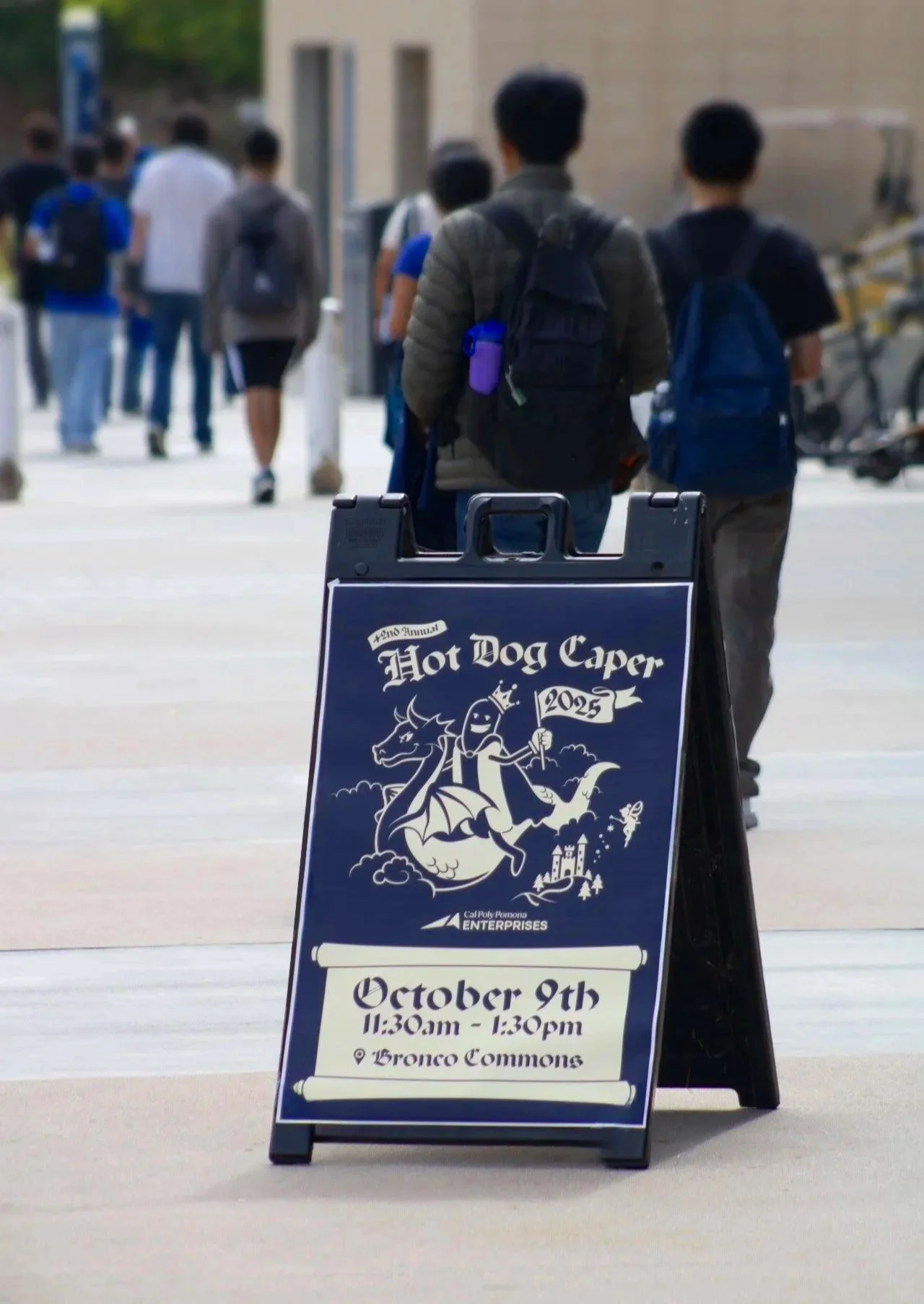 Event sign for the 2nd Annual Hot Dog Caper on October 9th from 11:30 am to 1:30 pm at Bronco Commons, featuring a cartoon hot dog character riding a dragon.