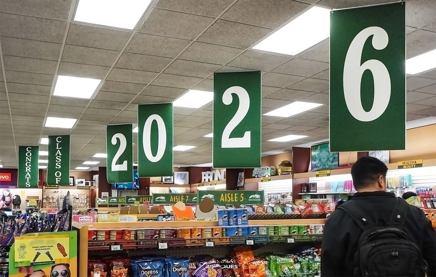 Grocery store aisle with large green signs displaying aisle numbers 6, 2, 2, 0, 2, 6 hanging from the ceiling. Shelves stocked with various snacks, beverages, and miscellaneous items. A person with a black backpack is browsing the aisle.