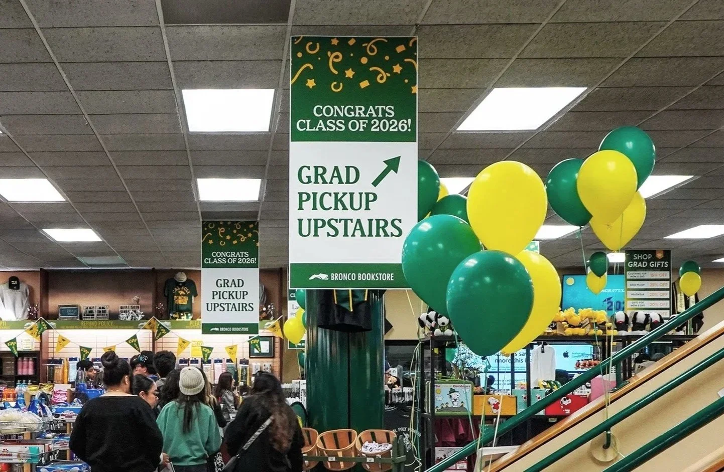 Group of people gathered inside Bronco Bookstore celebrating graduation, with green and yellow balloons, and signs indicating graduation class of 2026 and graduate pickup upstairs.
