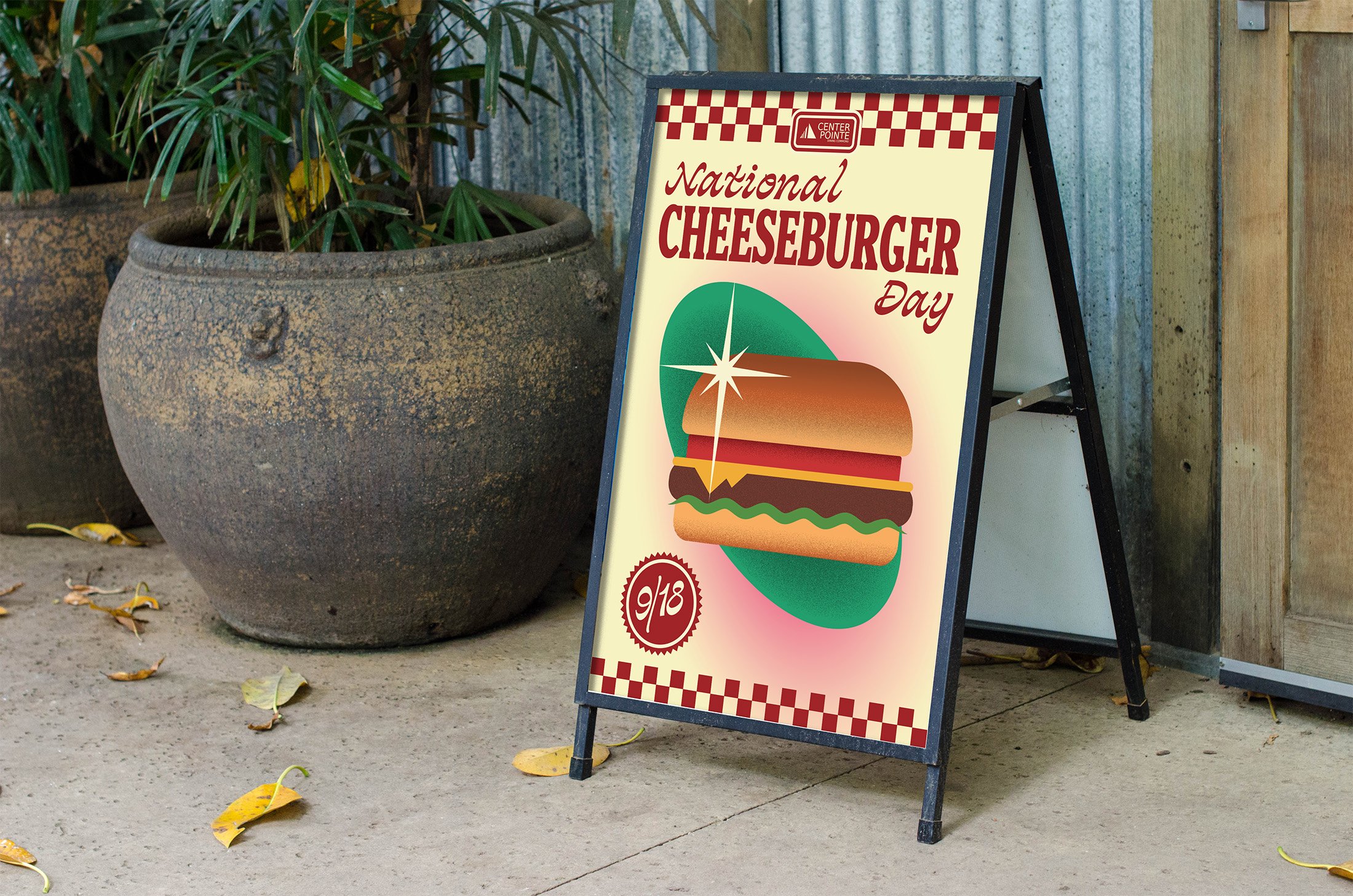A sandwich board sign advertising National Cheeseburger Day with a graphic of a cheeseburger, a checkered border, and red and cream colors, placed on a sidewalk next to a large plant in a pot and some fallen leaves.