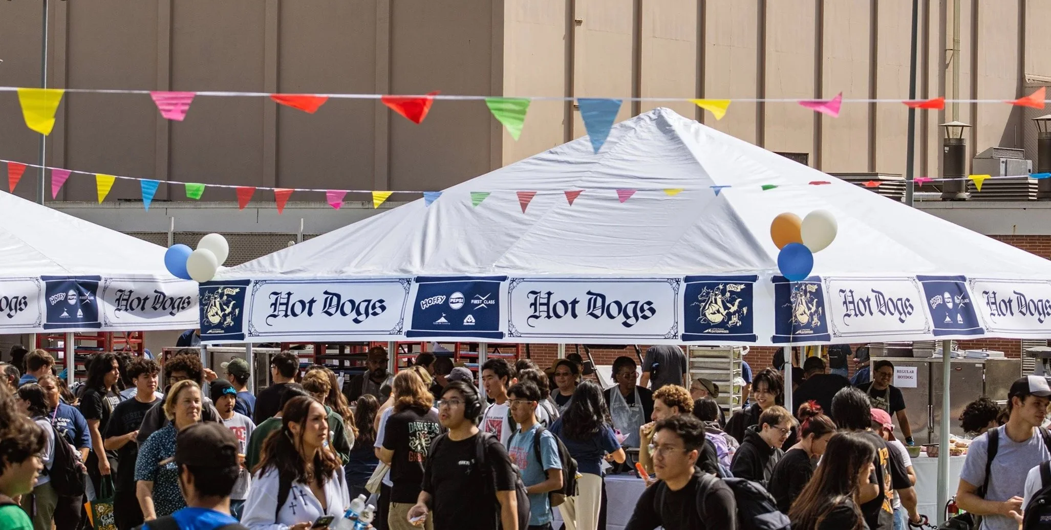Crowd of people at an outdoor festival or fair with a white tent selling hot dogs. Colorful pennant banners decorate the area. Many attendees are walking and talking.