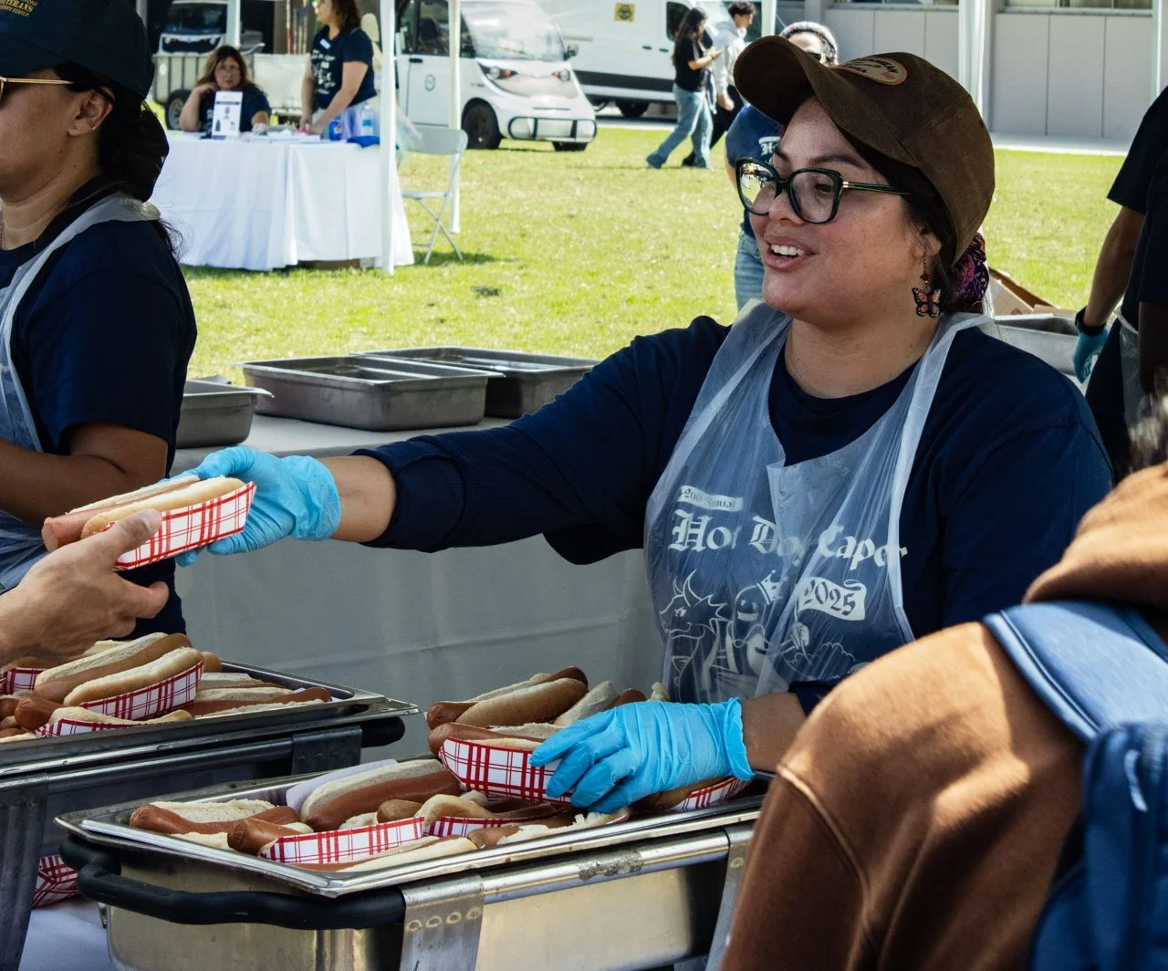 A woman wearing glasses, a brown cap, and a plastic apron is serving hot dogs at an outdoor event. She is handing a hot dog to a person in a brown jacket and blue glove. Several hot dogs are on trays in front of her, and there are other people and stalls in the background.
