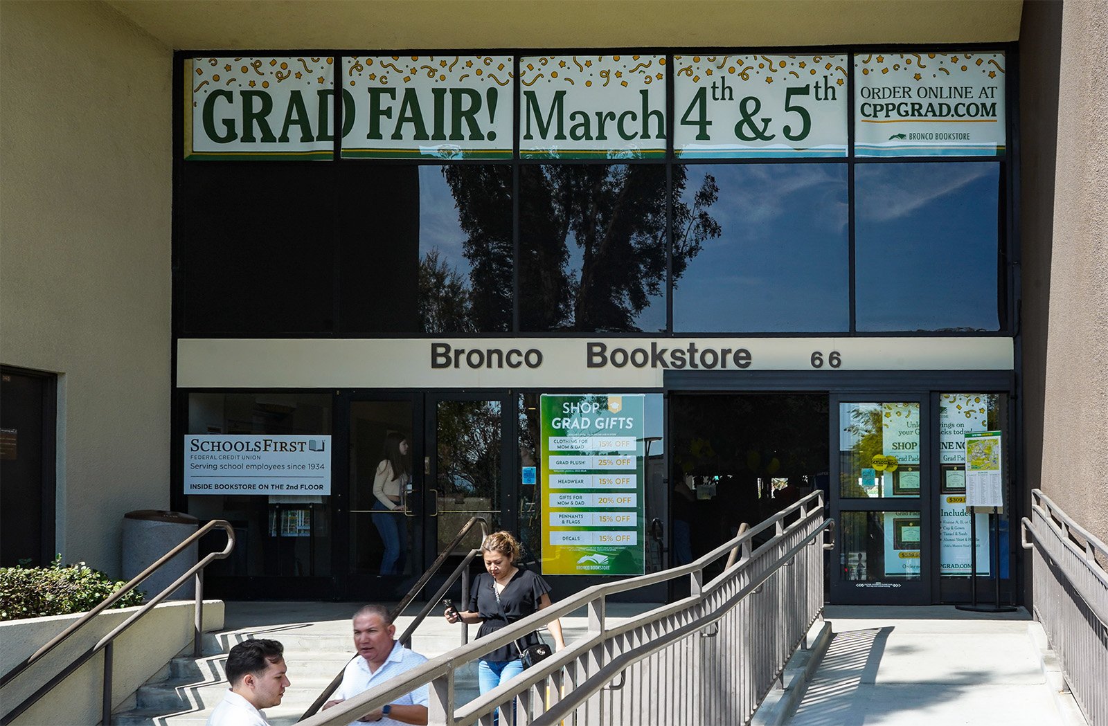 The entrance of Bronco Bookstore with a sign announcing a graduation fair on March 4th and 5th. There are people walking up and down the stairs outside the store.