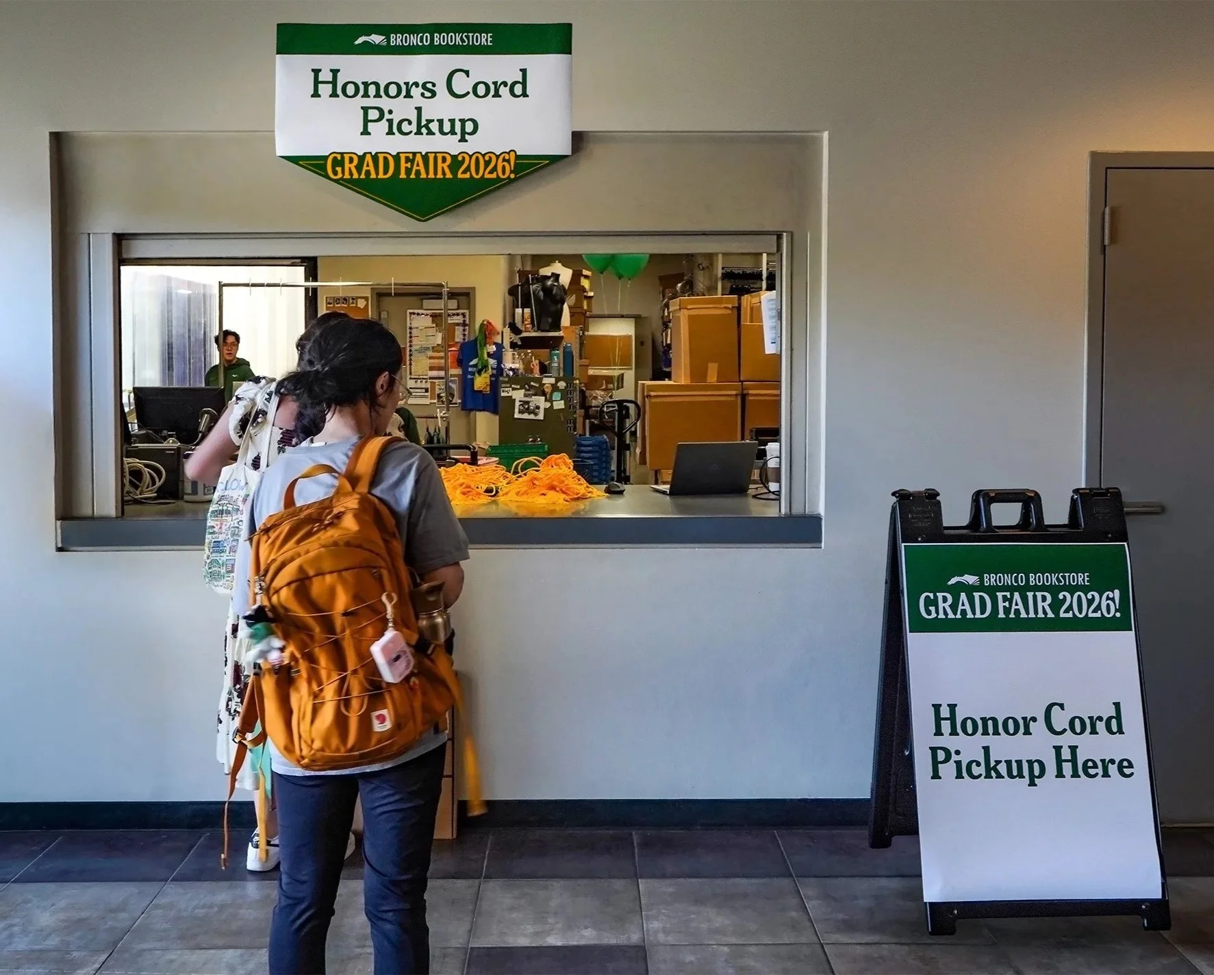 A student with a large orange backpack stands at a service window in a bookstore, waiting to pick up their honors cord for graduation. There is a sign above the window advertising honors cord pickup and a standing sign in front of the window with similar information for the graduation fair 2026.