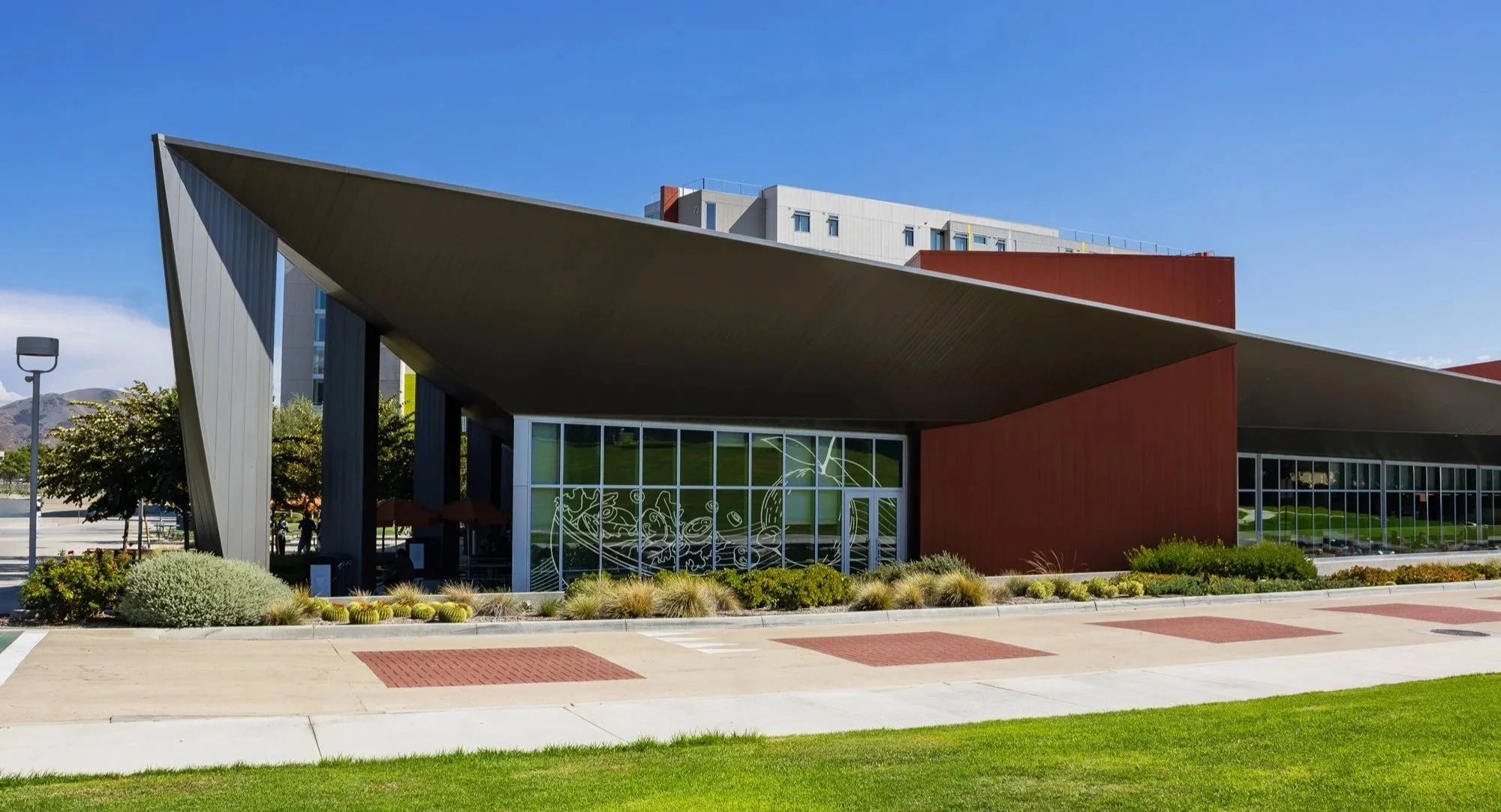 Modern building with angular roof and large glass windows, surrounded by landscaped plants and a clear blue sky.