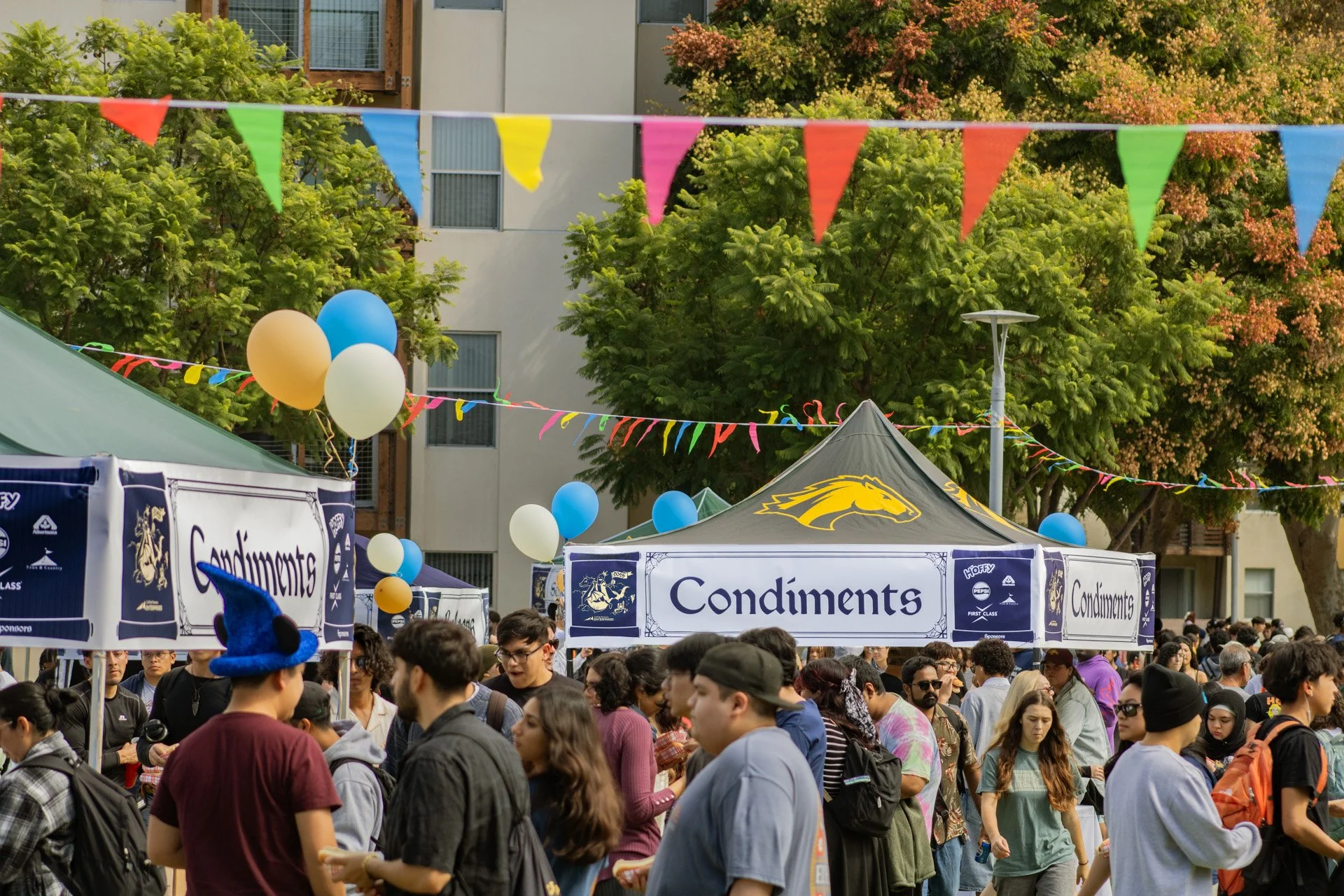 A crowded outdoor event with numerous people, several tents labeled 'Condiments,' colorful pennant banners overhead, and balloons, with trees and a building in the background.