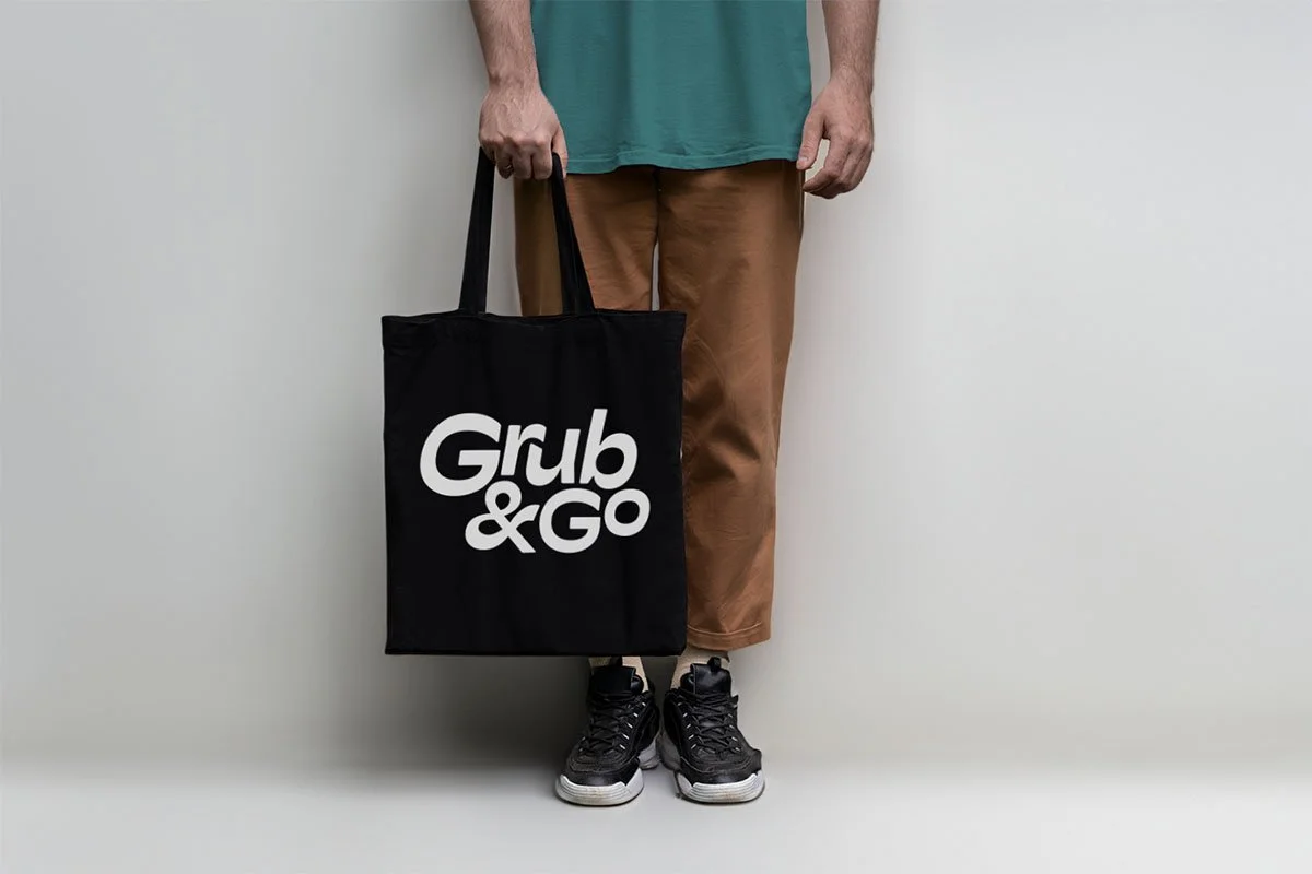 Person holding a black shopping bag with the words "Grub & Go" in white text, standing against a plain white wall, wearing brown pants, black sneakers, and a teal shirt.