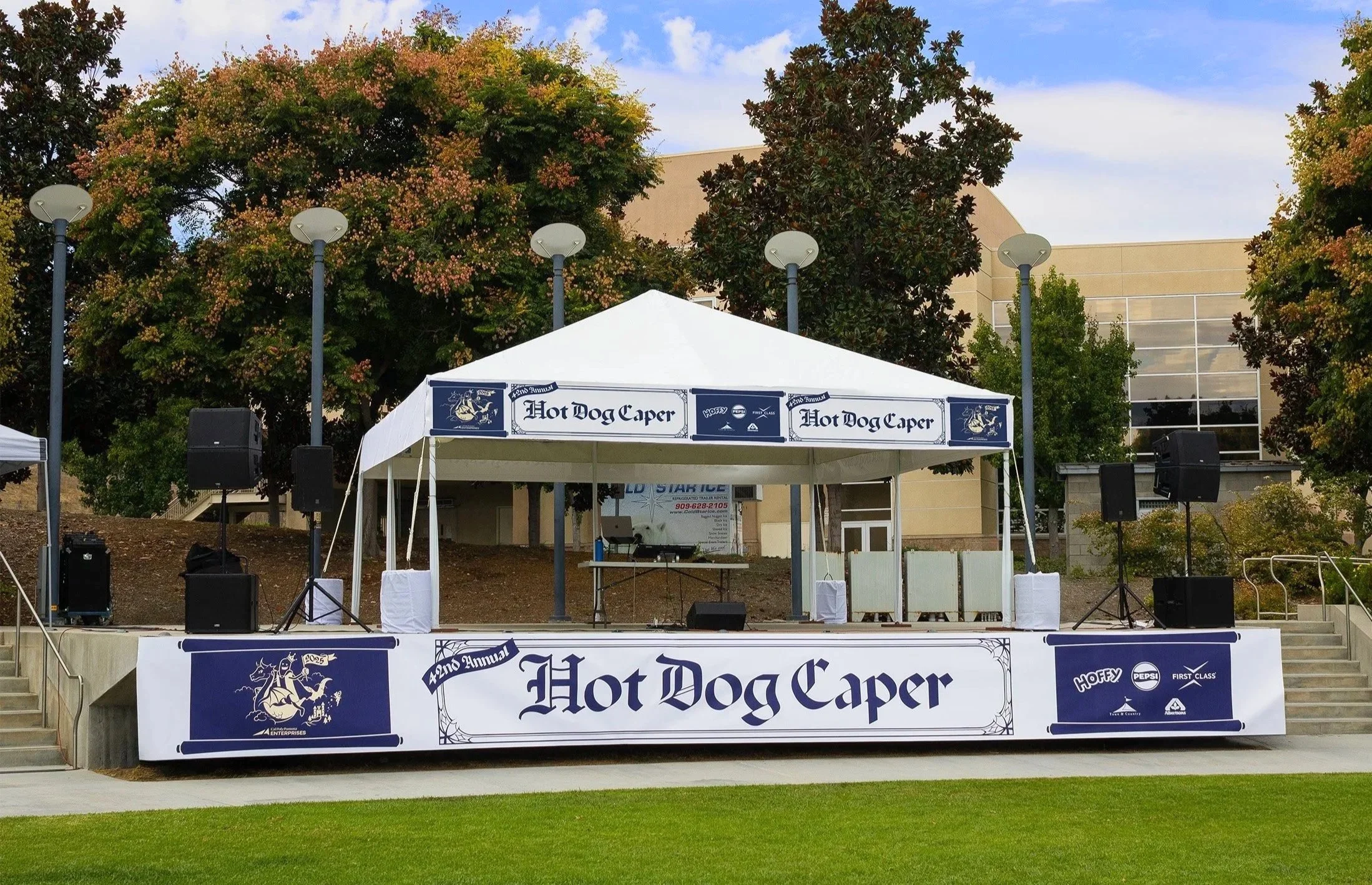 An outdoor stage setup with a white canopy tent displaying 'Hot Dog Caper, 42nd Annual,' with sound equipment and banners, and trees and a building in the background.