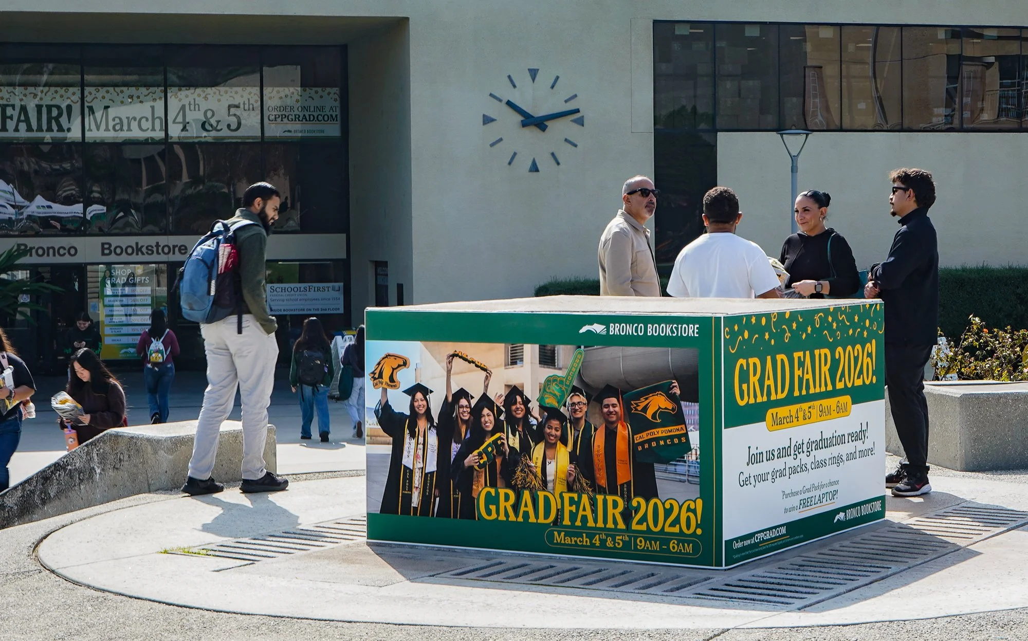 A group of diverse students and adults gathered outside a bookstore, with a sign announcing a graduation fair on March 4th and 5th, 2026, at Cal Poly Pomona. The scene includes a wall clock showing approximately 2:10, and the background features large windows and an urban setting.