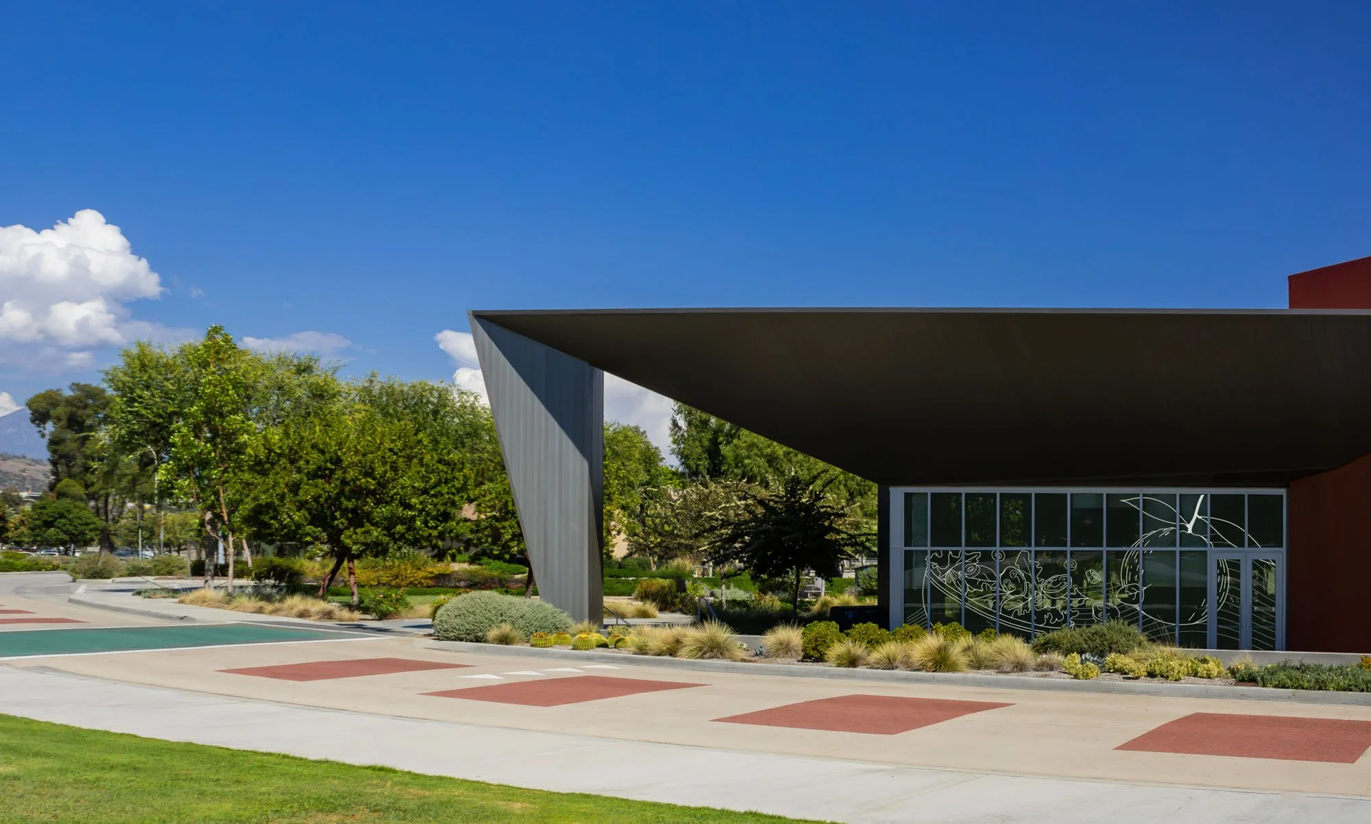 Modern building with angular roof overhang, large glass window with abstract art, surrounded by landscaped trees and bushes under a bright blue sky.