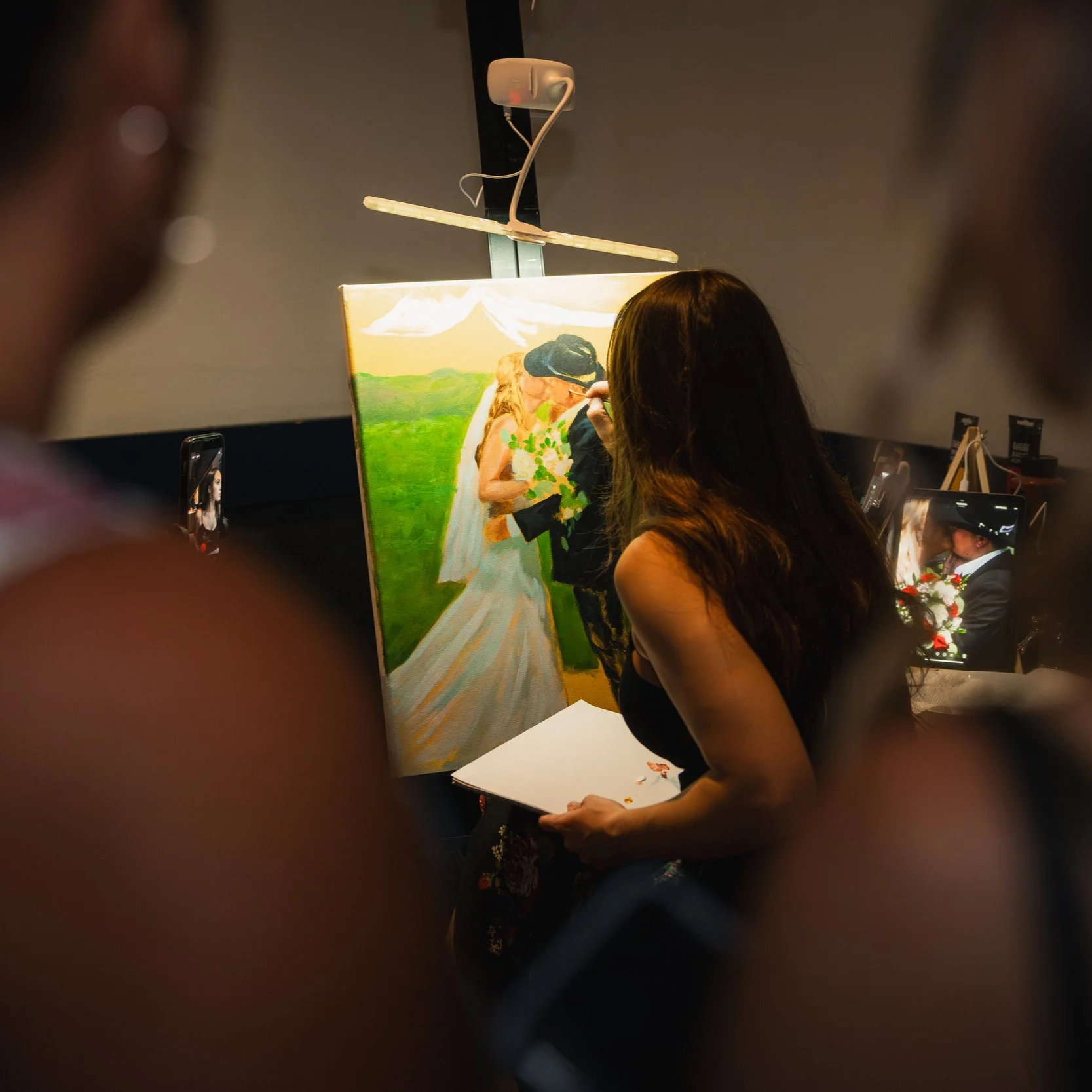 A woman painting a portrait of a couple kissing outdoors, with a green landscape and mountains in the background, under a cloudy sky.