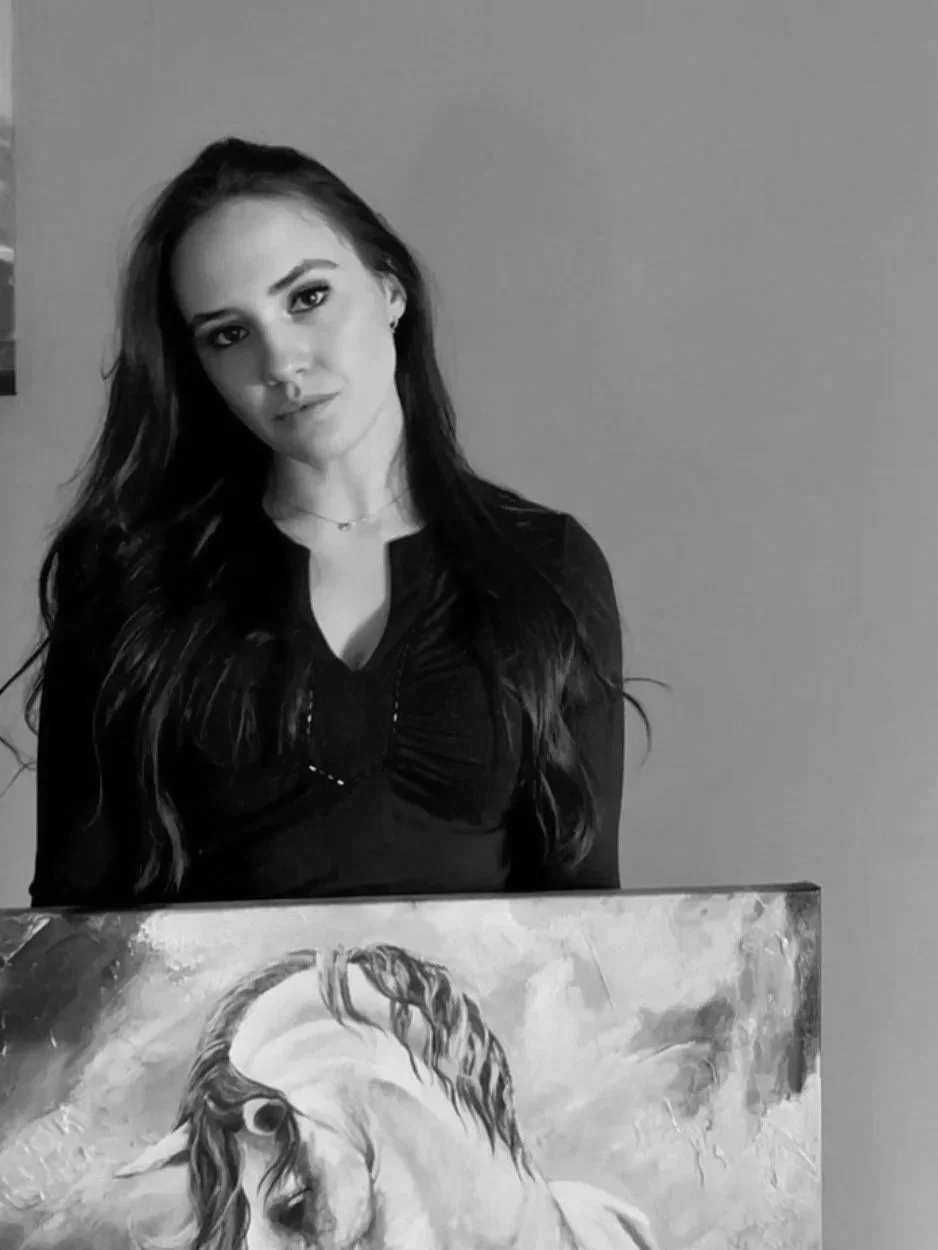 A young woman with long dark hair and light skin standing next to a painting of a unicorn with a flowing mane, set against a plain wall.