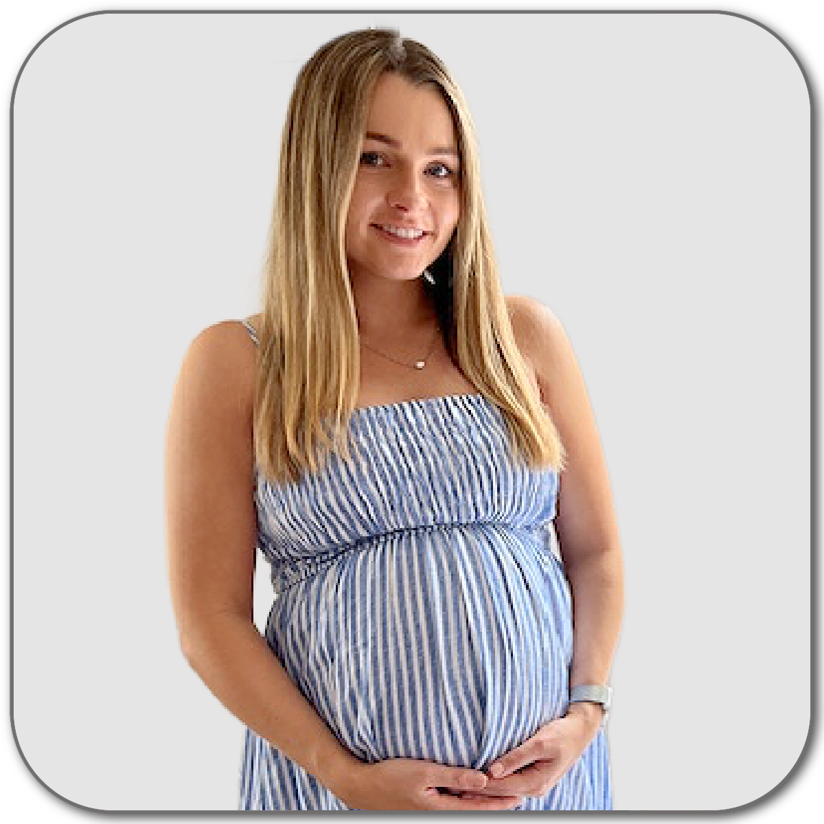 A pregnant woman with long blonde hair smiling, wearing a blue and white striped sleeveless dress, standing against a plain light gray background.