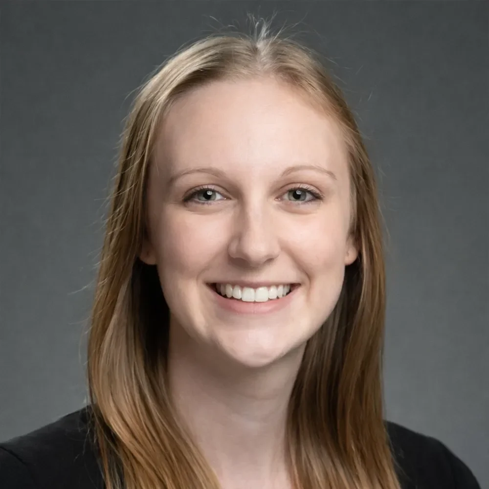 A young woman with long red hair smiling at the camera, wearing a black top, against a plain gray background.