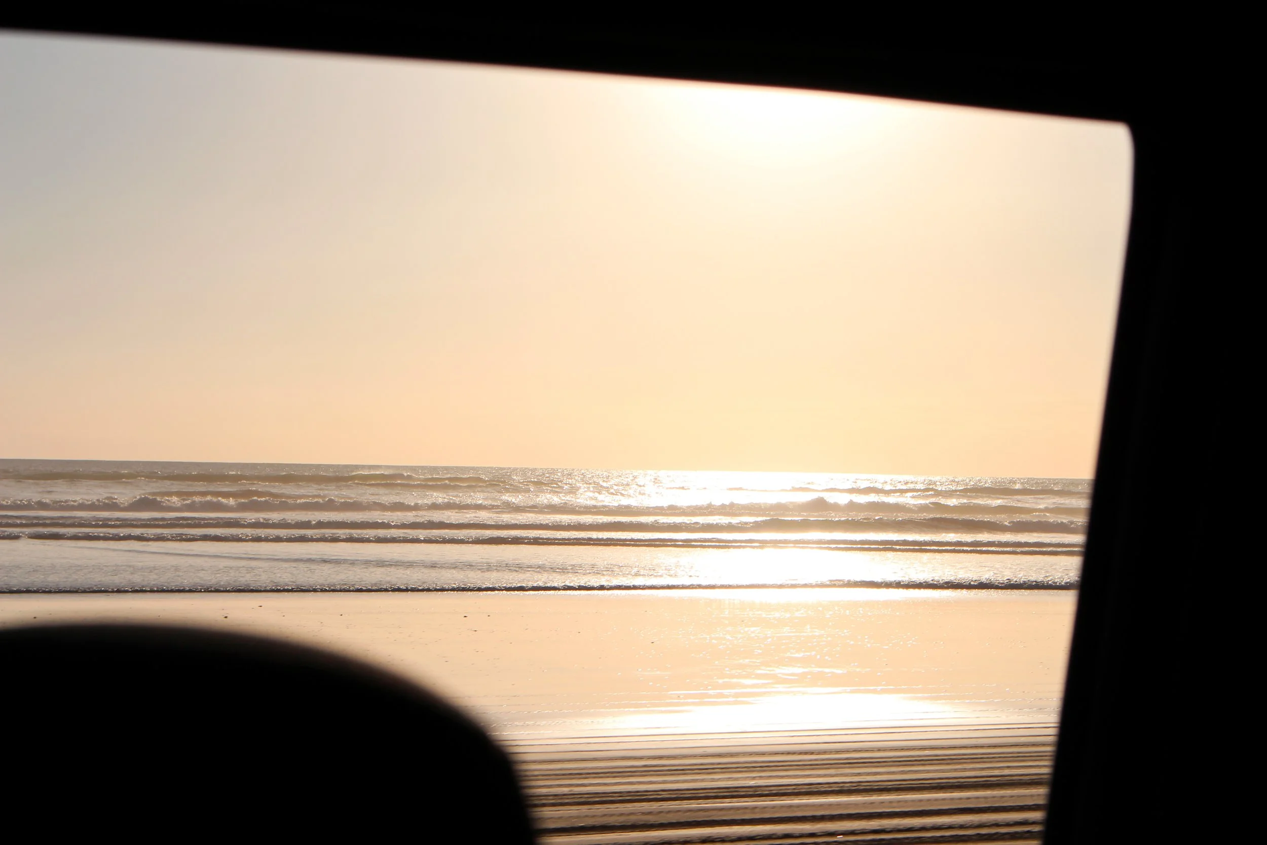View of the ocean and sunset through a window with black blinds partially open.