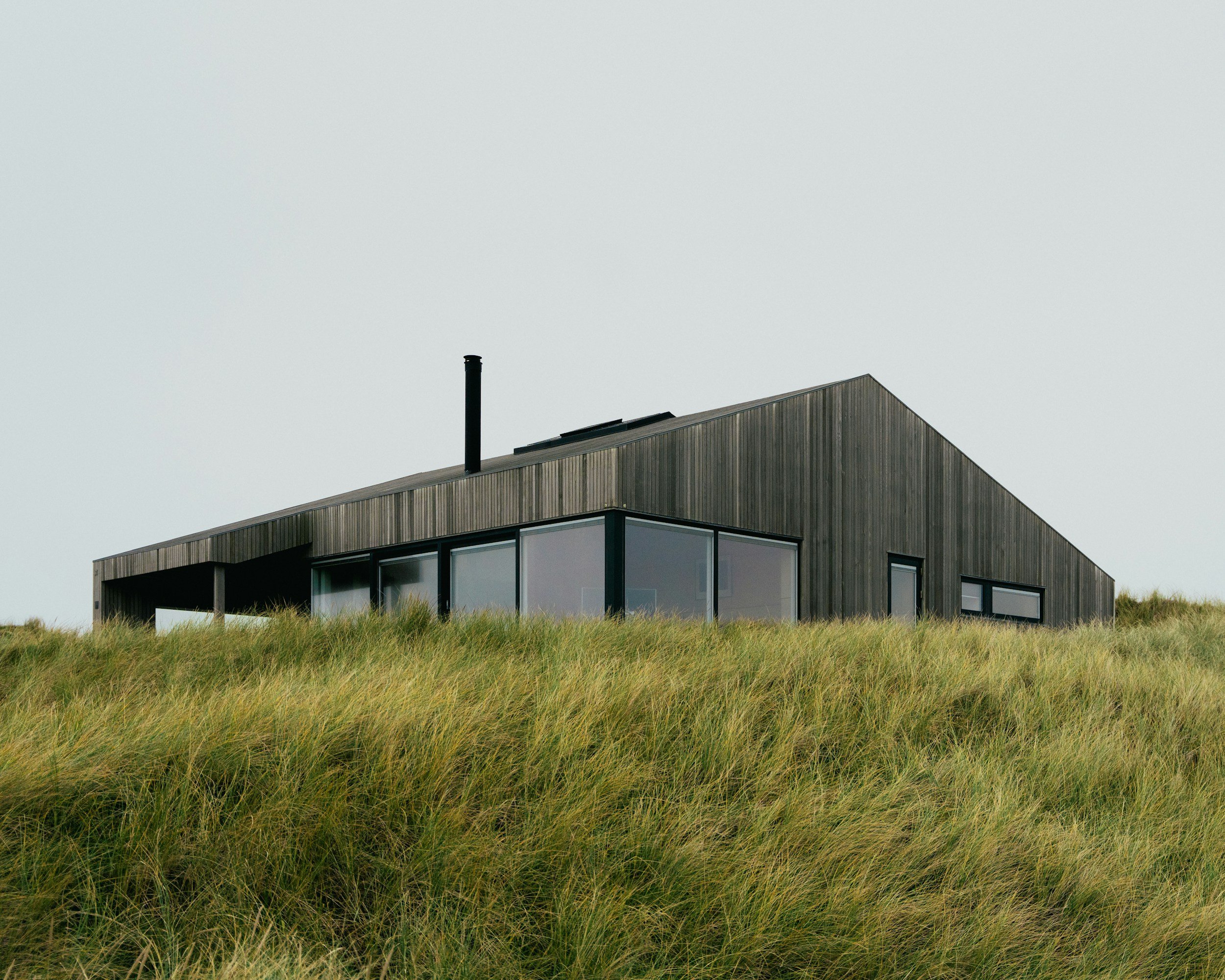 Modern building with wooden exterior and large glass windows, surrounded by tall grass, under an overcast sky.