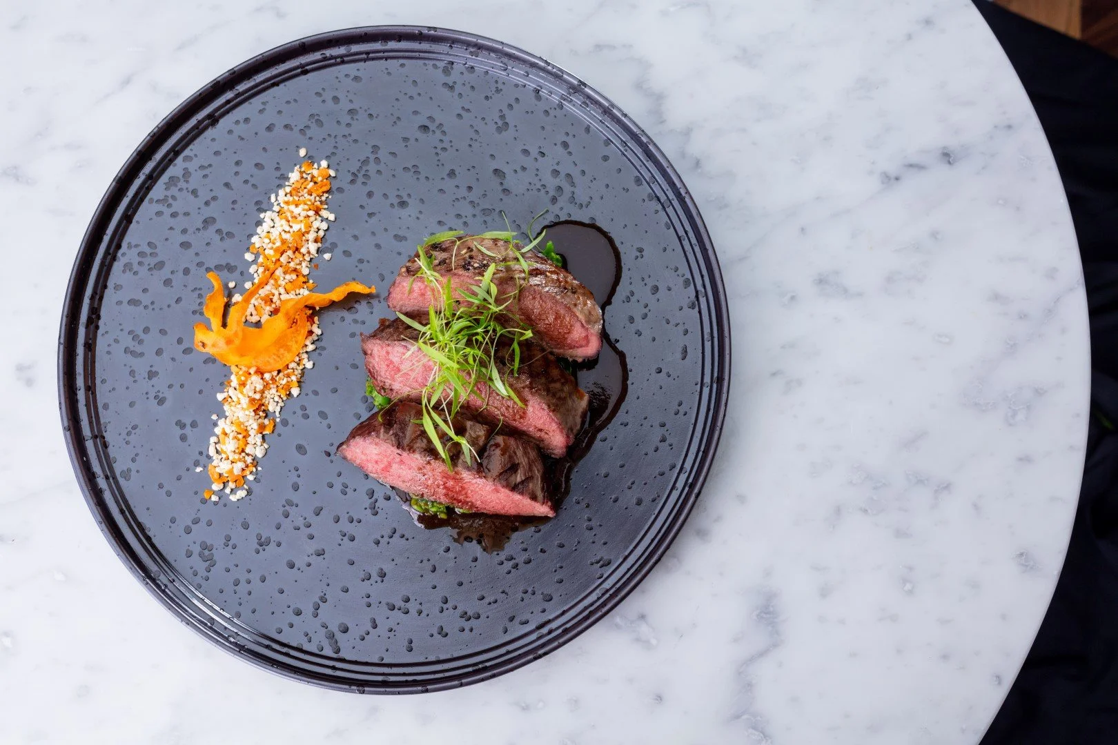 Sliced medium-rare steak garnished with greens on a black plate, accompanied by a smear of orange puree and a sprinkle of grains, served on a white marble surface.