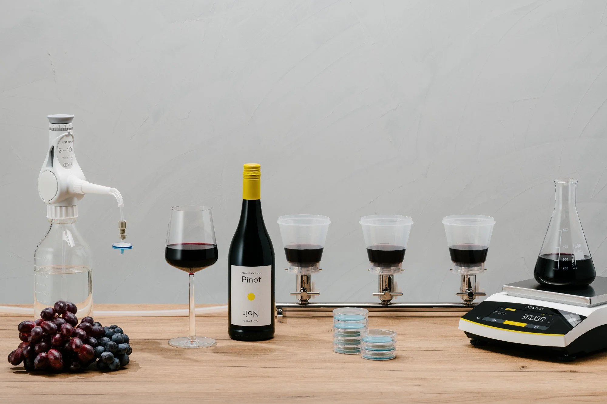Laboratorio de vino con una botella, copas con vino, tinajas, y utensilios de medición sobre una mesa de madera contra una pared gris.