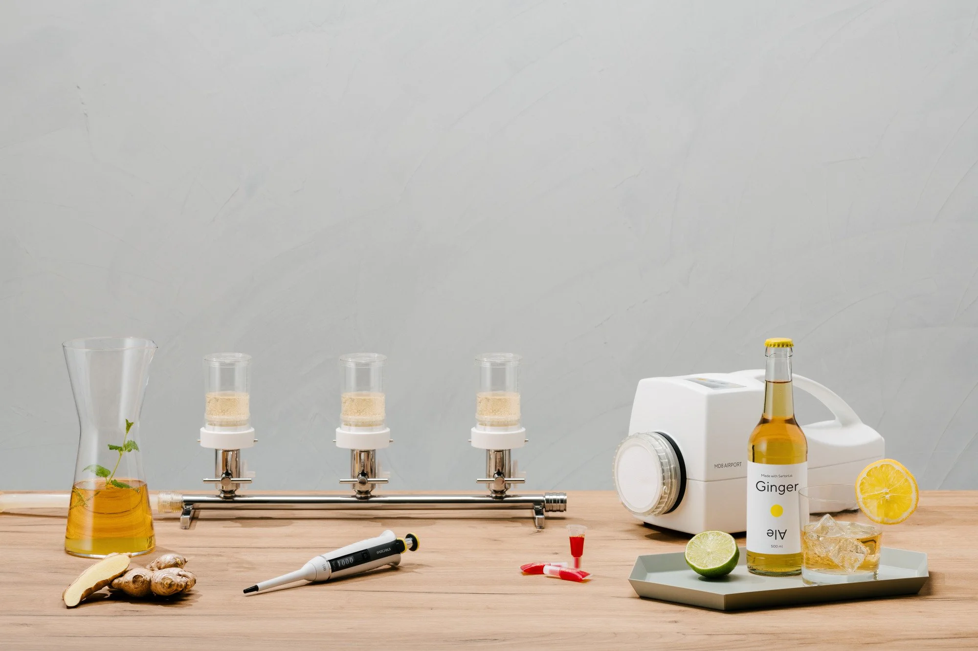 Mesa de laboratorio con frascos dispensadores, una garra con hojas, un vaso con raíz, jugo de jengibre, limones, una máquina de jugo, un bolígrafo, y tubos de ensayo con líquidos y etiquetas.