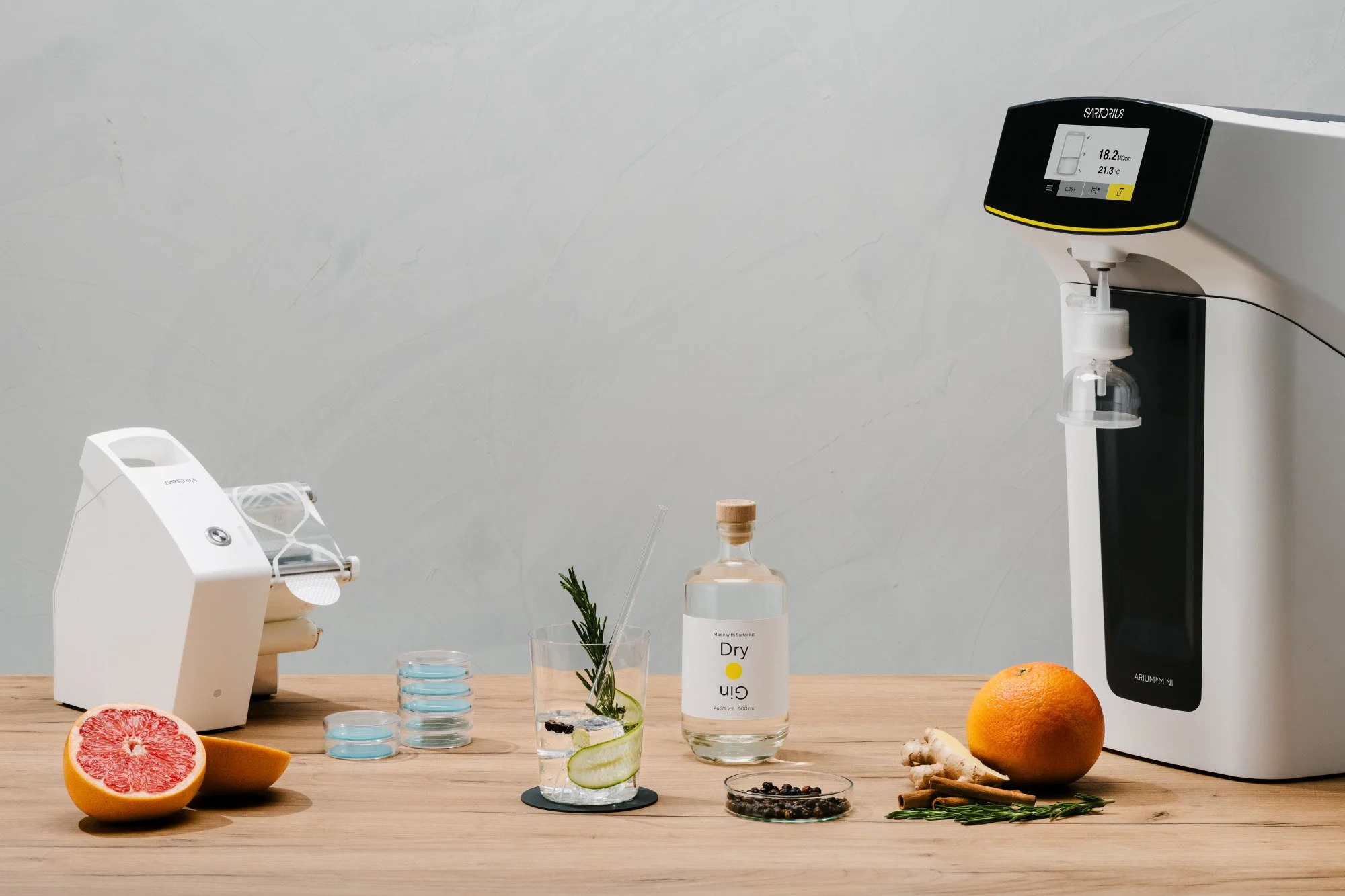 Una mesa de madera con cítricos, jengibre, hierbas, vasos de agua, y un equipo de laboratorio para experimentos culinarios, además de un monitor digital.