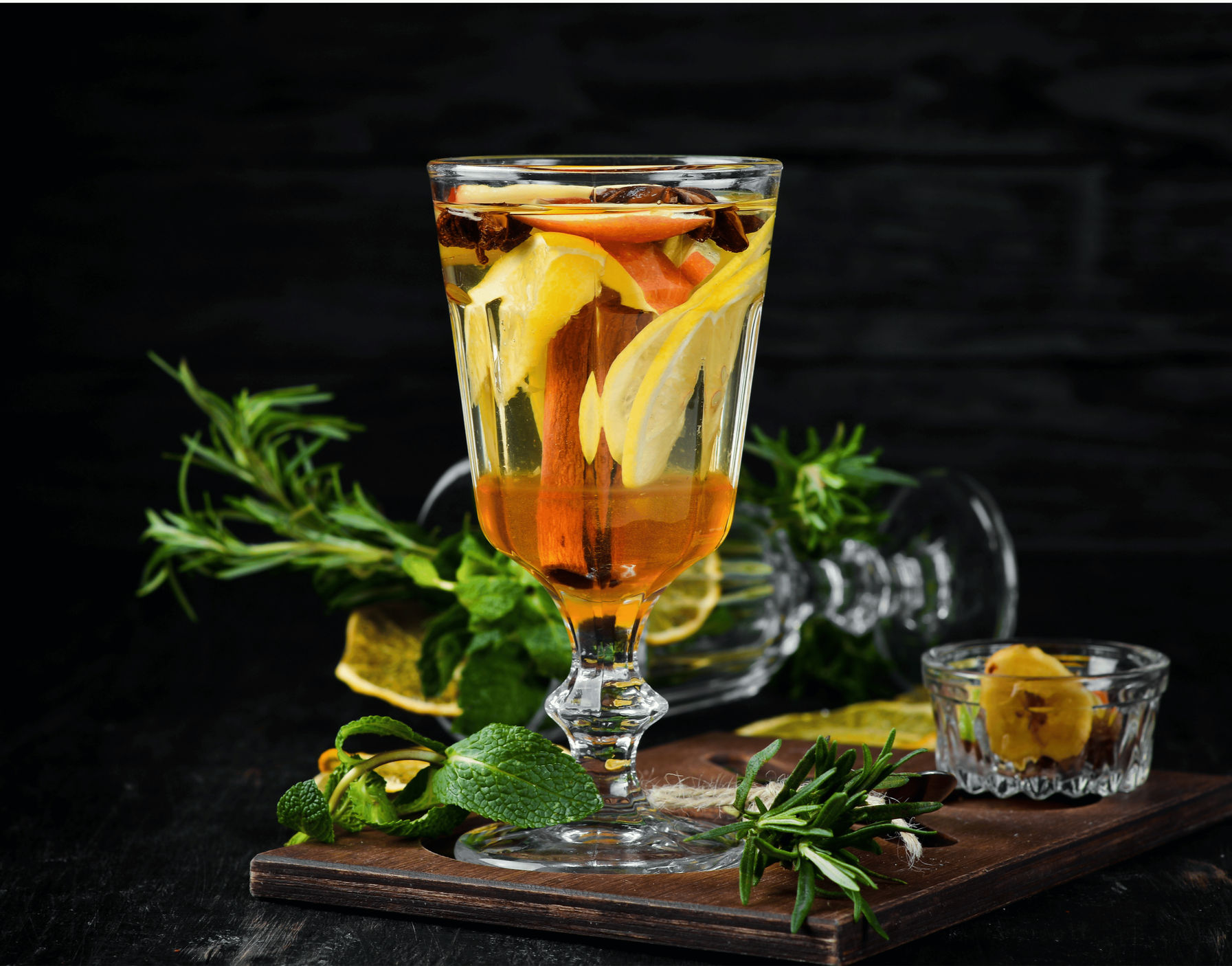 Vaso con infusión de limón, naranja, canela, y bayas en un fondo negro, decorado con hierbas y rodajas de cítricos.