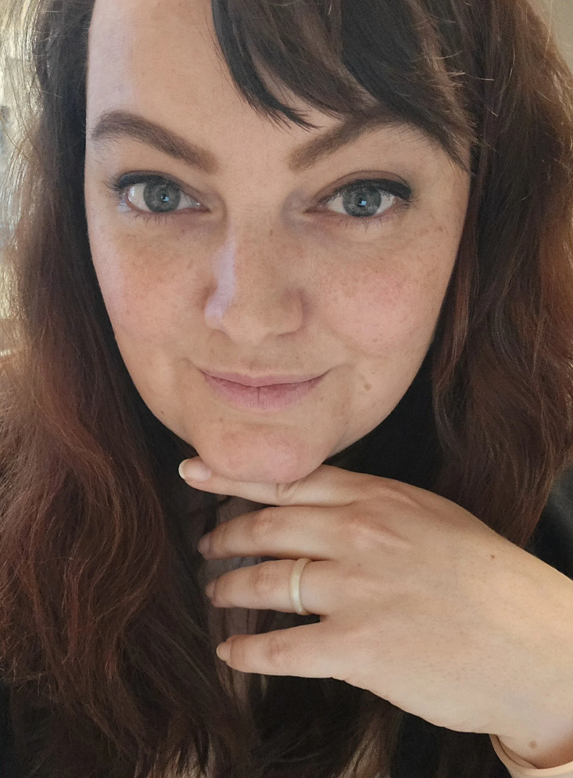 A close-up of a woman with long reddish-brown hair, blue eyes, and light skin, resting her hand near her chin with a gold ring on her finger.