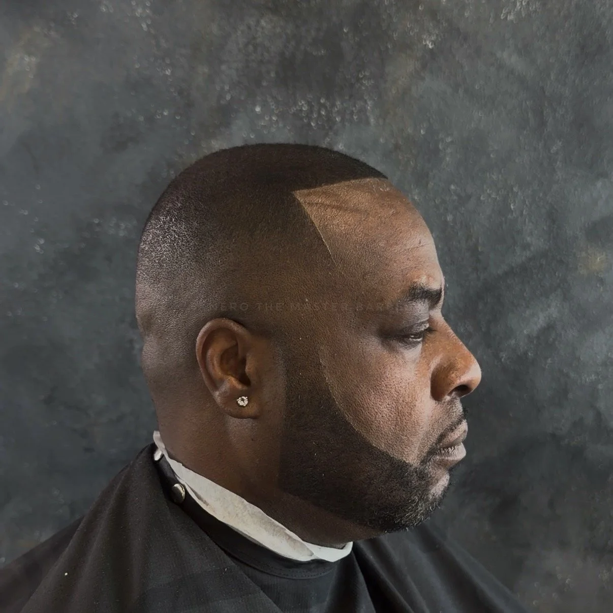 Side profile of a man with a fresh, close-cropped haircut featuring a geometric design, wearing a black barber cape and a white shirt with a black choker.