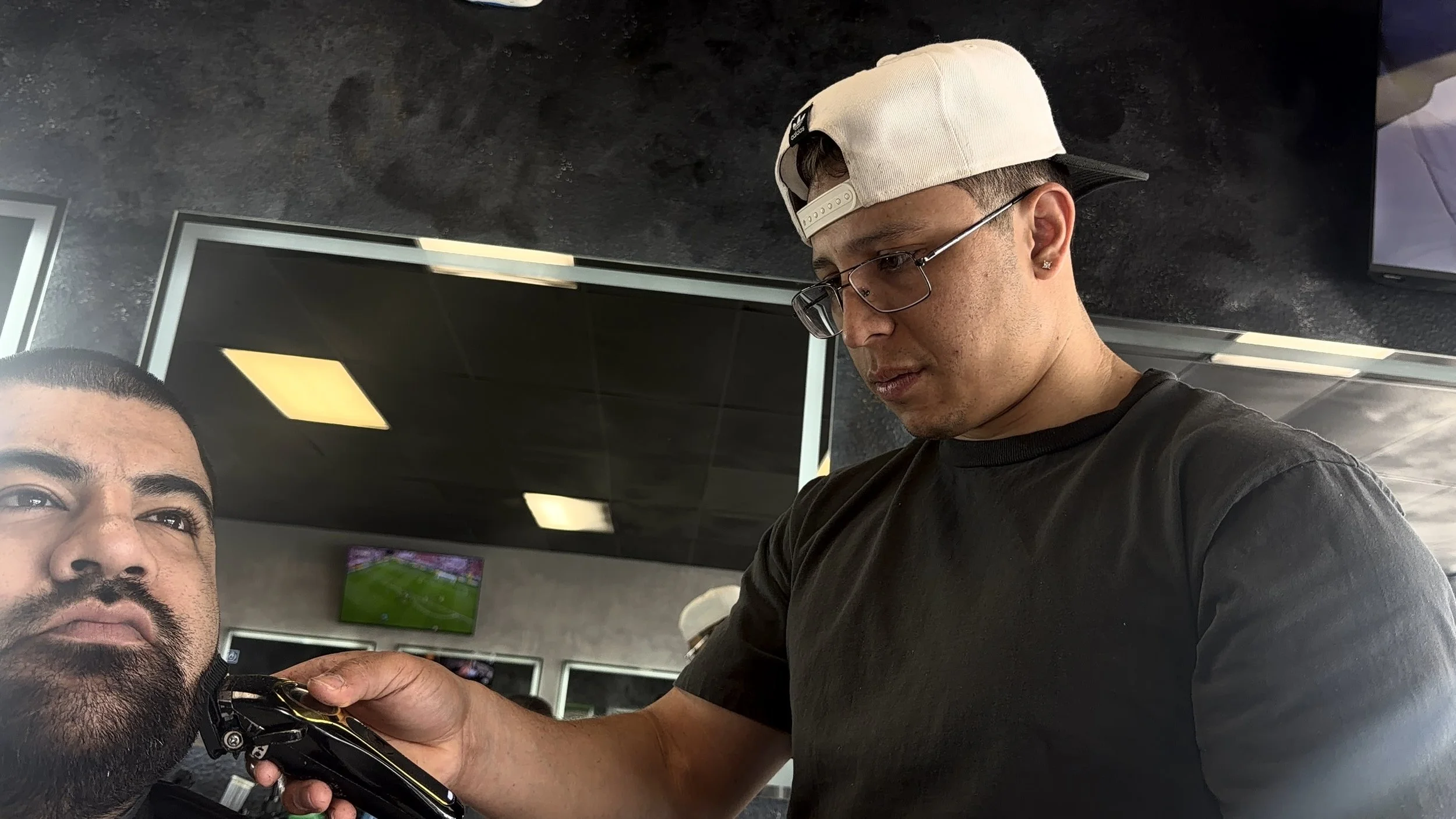 A person with glasses and a white backwards cap using electric shaver on another person's beard, in a room with black ceiling tiles and a TV screen in the background.