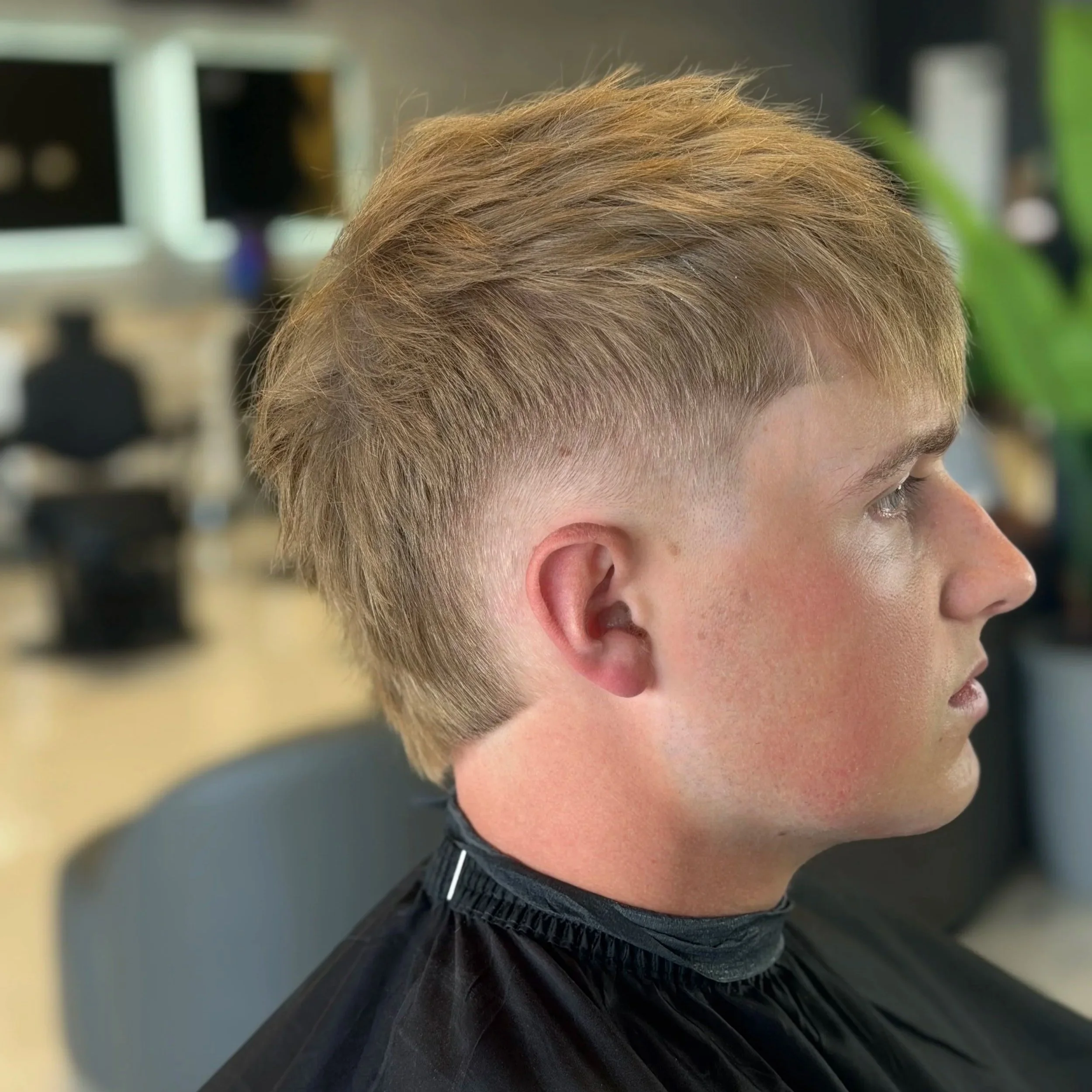 Close-up of a person's head with styled blond hair, wearing a black barber cape, in a barbershop setting.