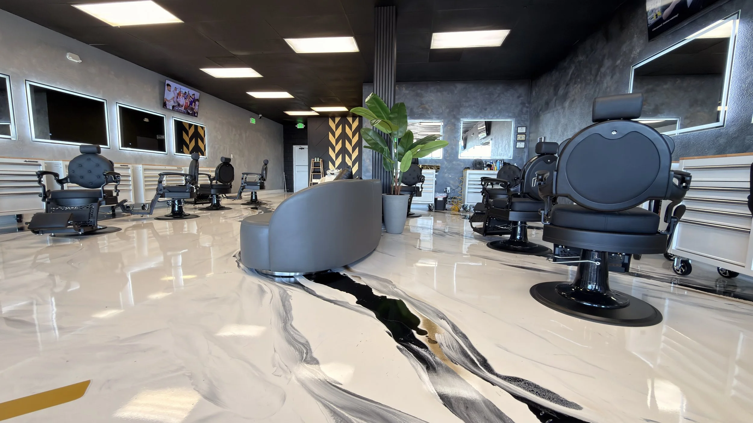 Interior of a modern barbershop with several black barber chairs, a large potted plant, mirrors on the walls, and a marble floor with black and white veining.