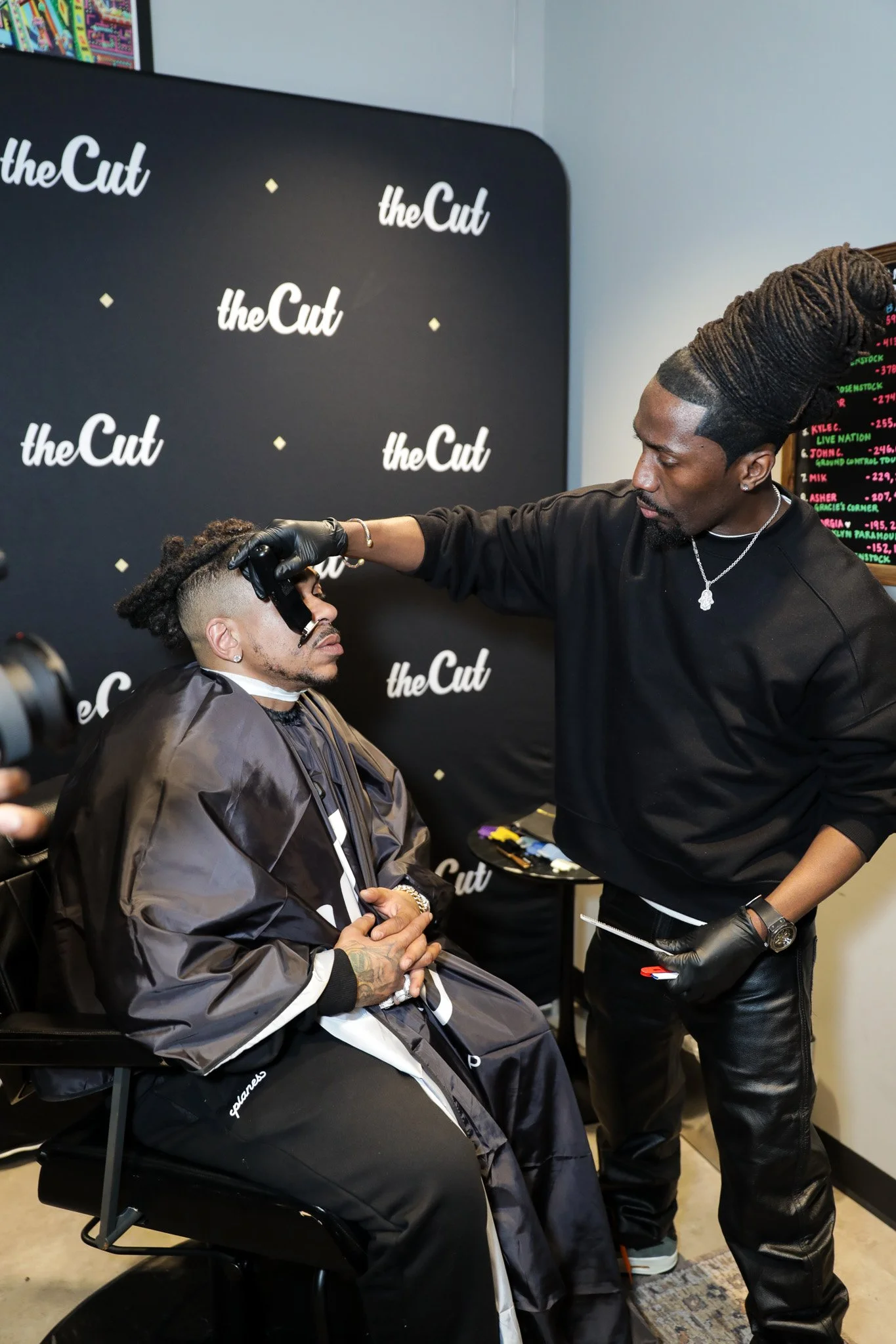 A man sitting in a barber chair getting a haircut from a barber in a salon with 'theCut' logos on the wall behind them.