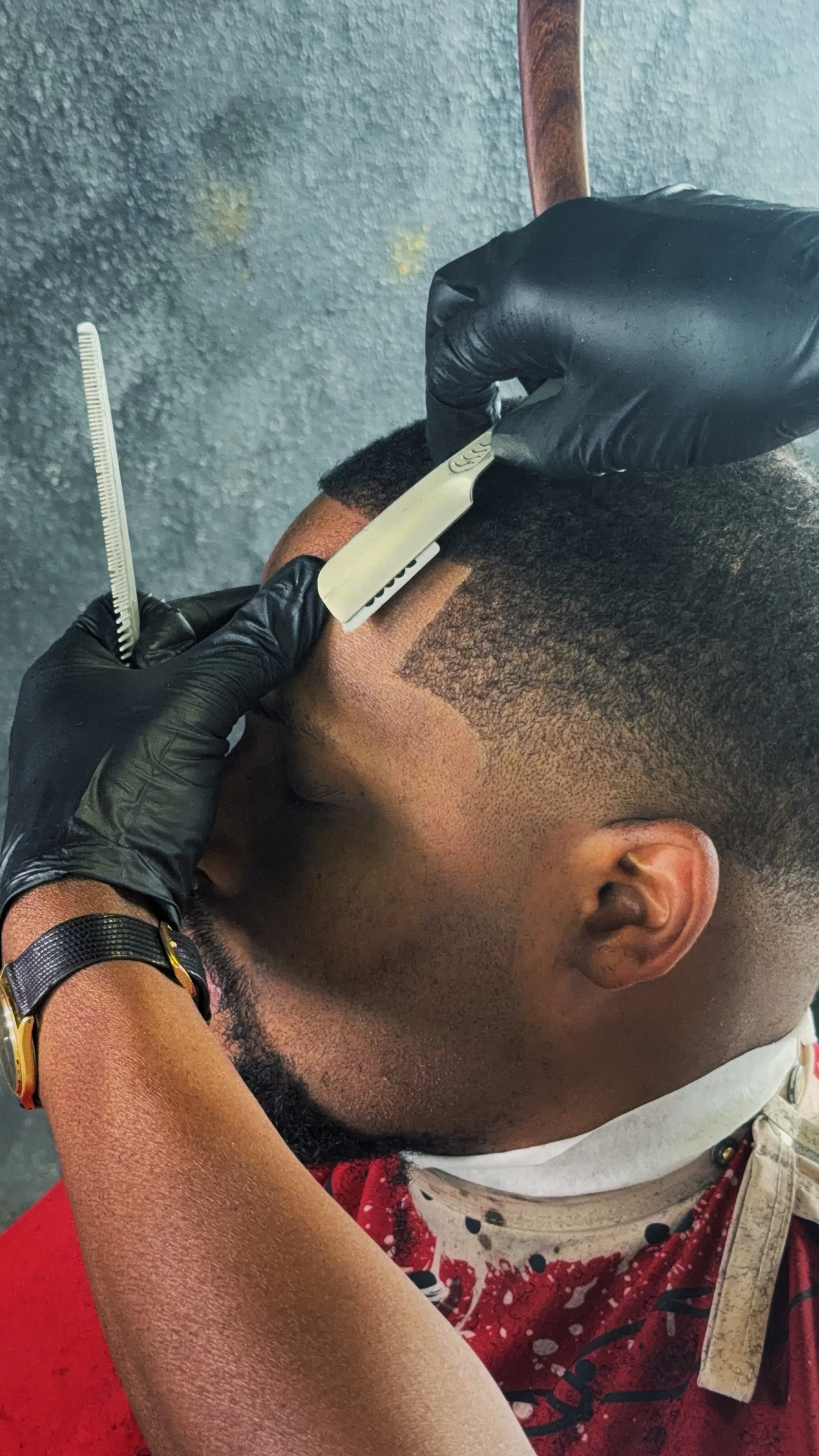 A person getting a haircut in a barbershop, with a barber using clippers on the person's hair and holding a comb, both wearing black gloves.