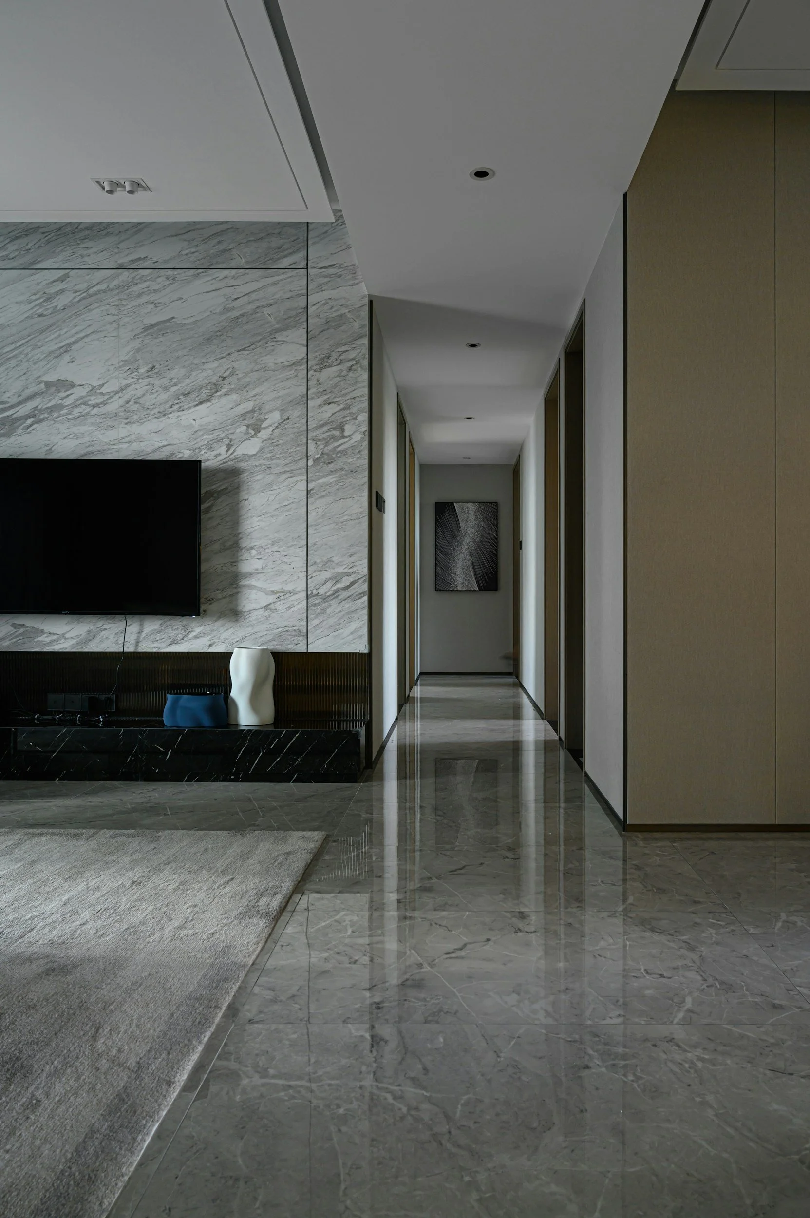 Modern hallway with marble flooring and walls, featuring a hallway with multiple doors, a TV on the left wall, decorative vases, and minimalist artwork.