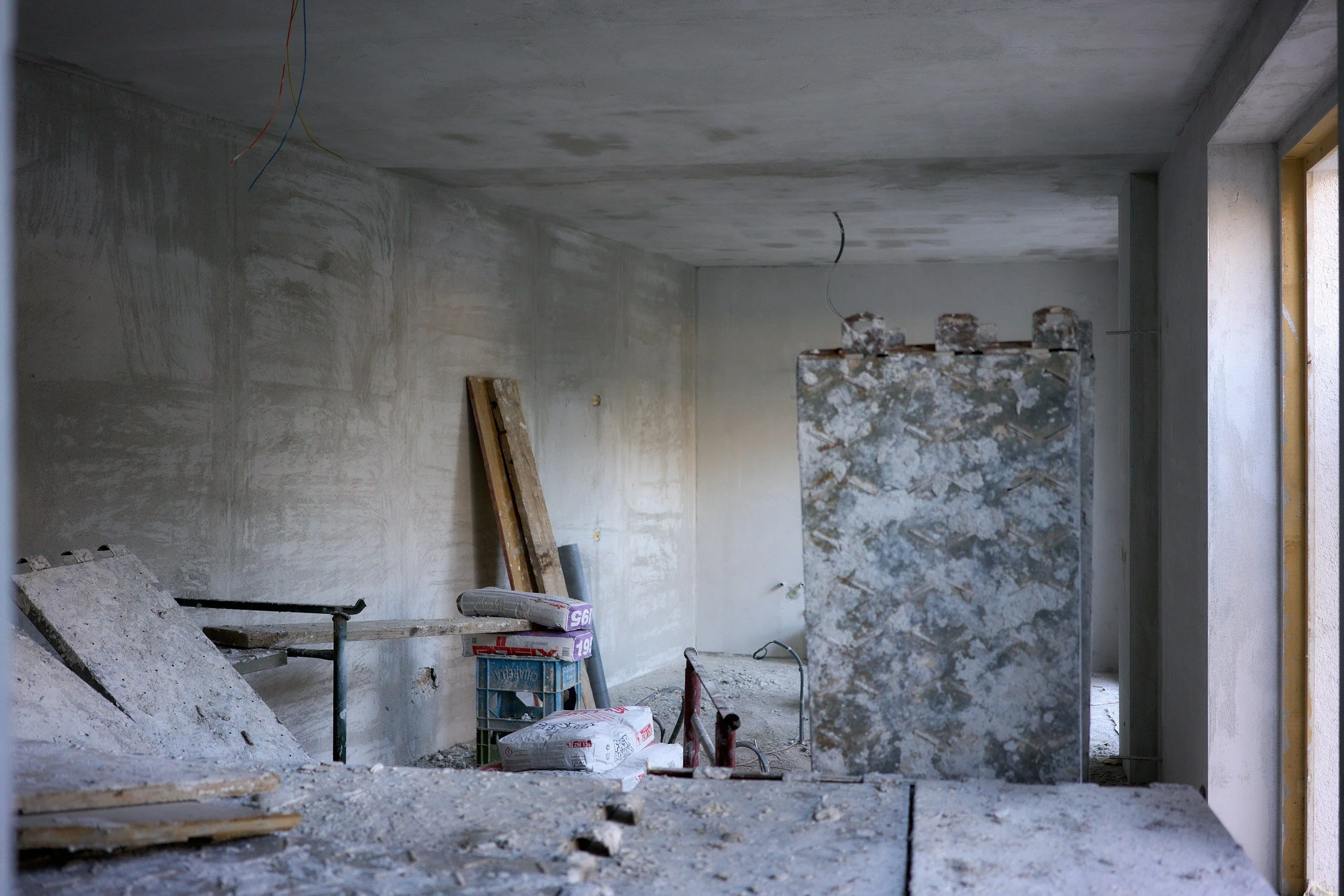 An unfinished room under construction with plastered walls, exposed wiring from the ceiling, construction materials, and tools scattered on the floor.