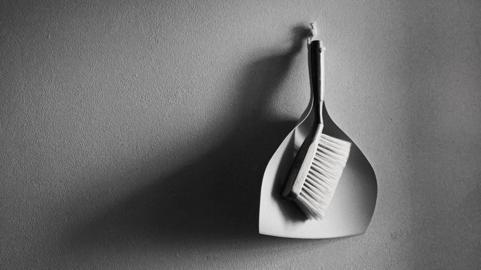 A dustpan with a cleaning brush resting inside it, placed on a textured surface, with shadows cast to the left side.