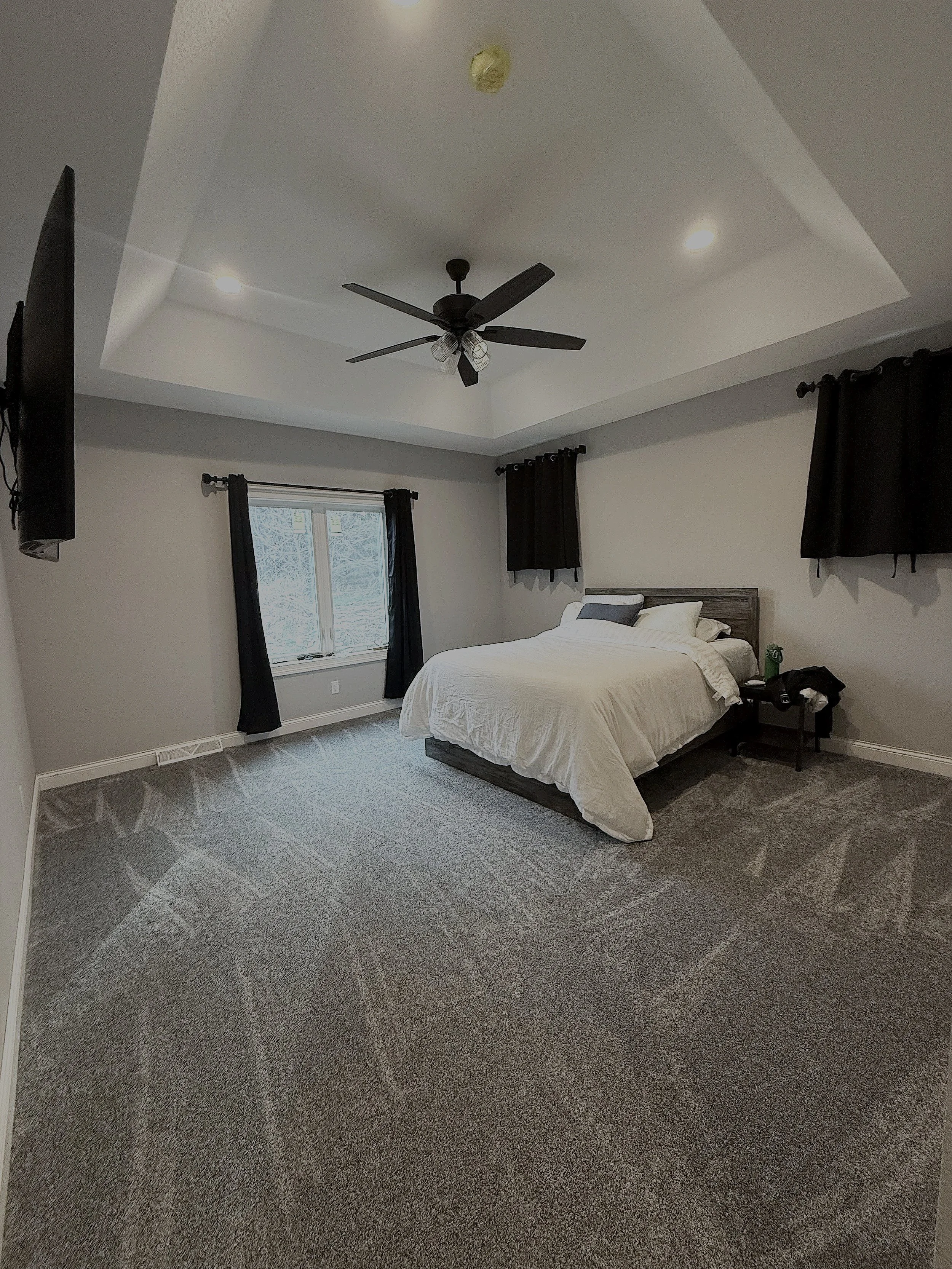 A bedroom with a bed, window with black curtains, ceiling fan, and wall-mounted TV.
