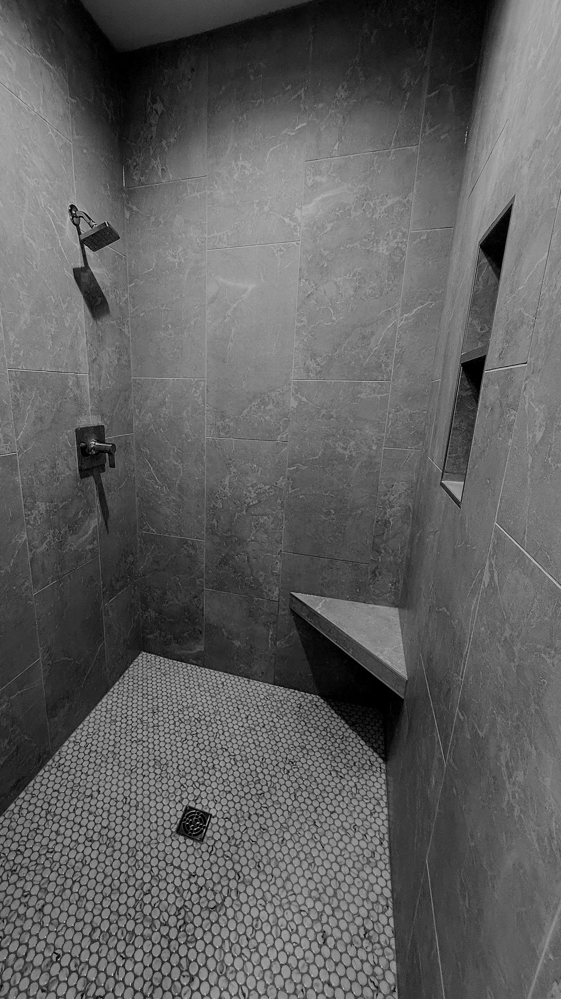 A modern shower with gray marble walls, a black shower head, a hand-held shower attachment, a built-in wall niche, a bench, and small hexagonal white tile flooring with a drain.