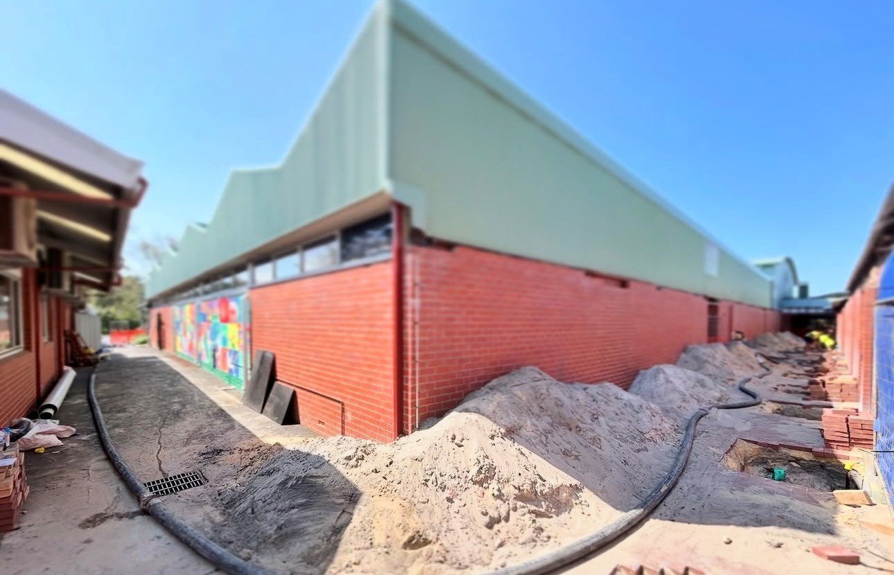 Construction site with piles of dirt, construction pipes, and a building with a red brick wall and a green siding exterior, next to a narrow pathway.