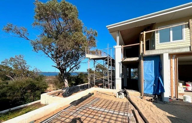 Construction site of a house with scaffolding, unfinished concrete foundation, and partially built exterior walls, with a clear blue sky and trees in the background.