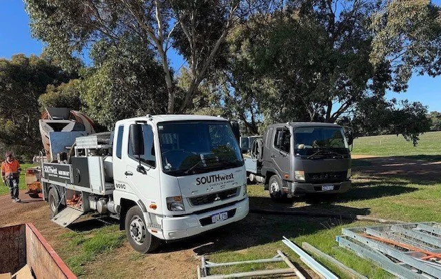 Two trucks parked on grass in a park-like setting, with trees and open space in the background. One is a white utility truck, and the other is a dark gray or black flatbed truck. Construction materials and tools are visible nearby.
