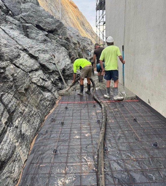 Construction workers pouring concrete on a rebar grid floor, with rock face on one side and scaffolding on the other.