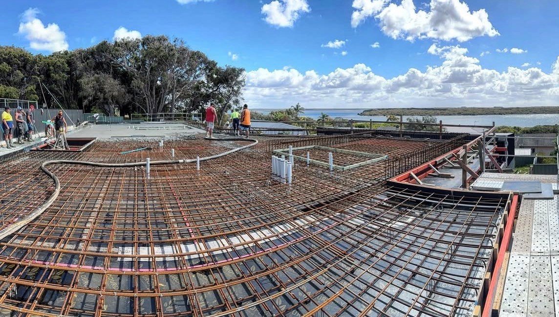 Construction workers on a building site with steel rebar framework, overlooking a body of water and trees under a partly cloudy sky.